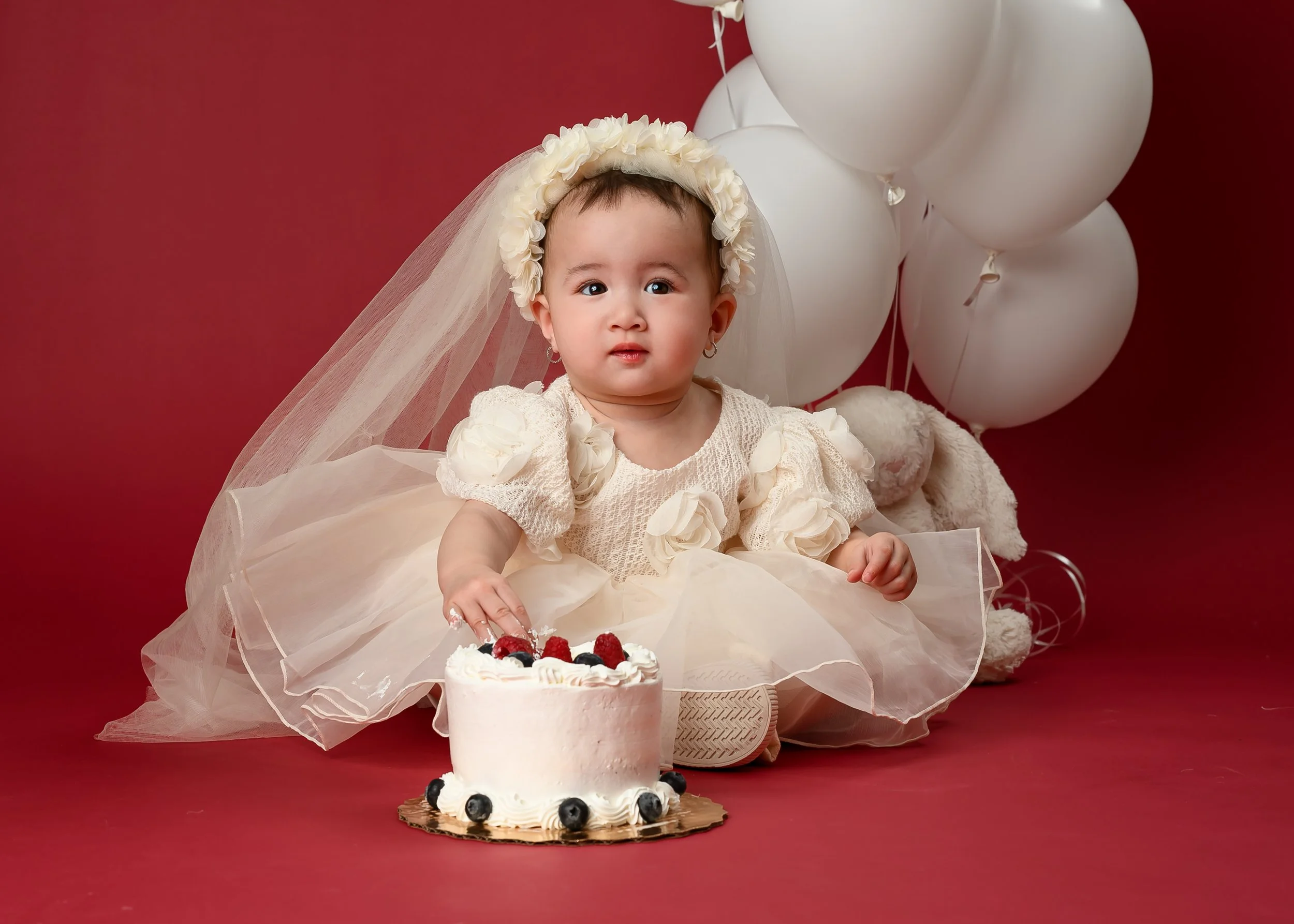 Baby C sitting in a cream dress and floral headband, gently touching her first birthday cake with white balloons behind her.