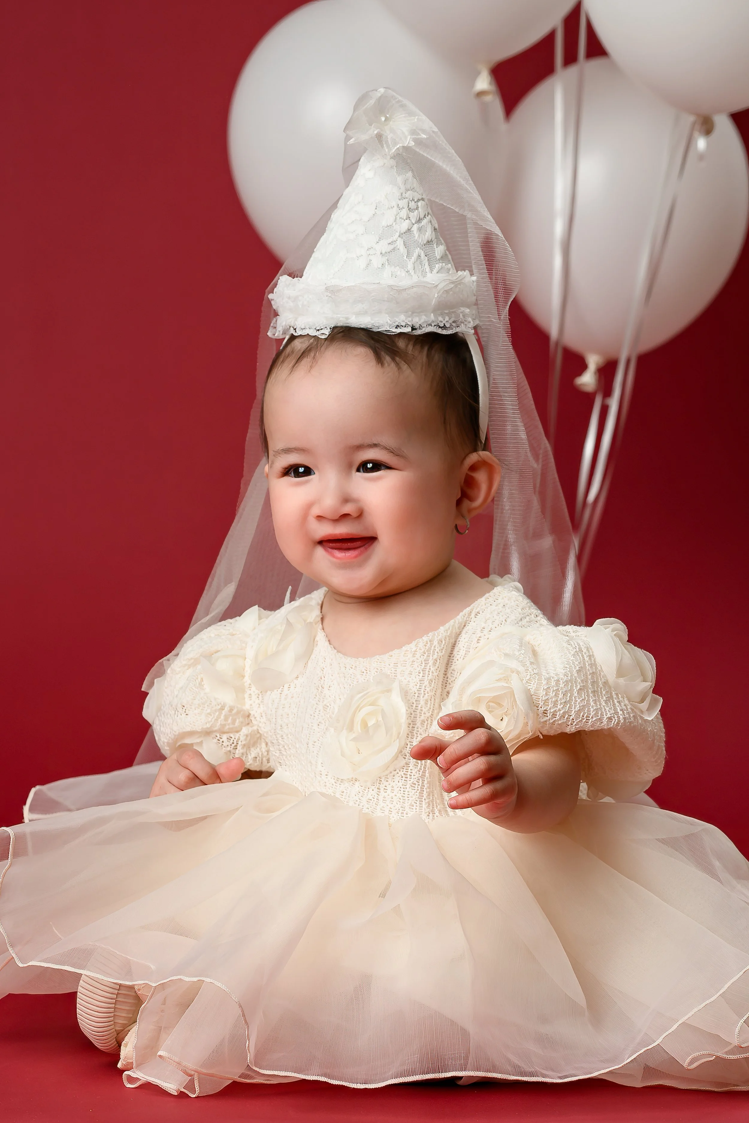 One-year-old baby girl wearing a cream dress and lace cone hat, seated in front of white balloons during her Sweet Celebrations birthday session.