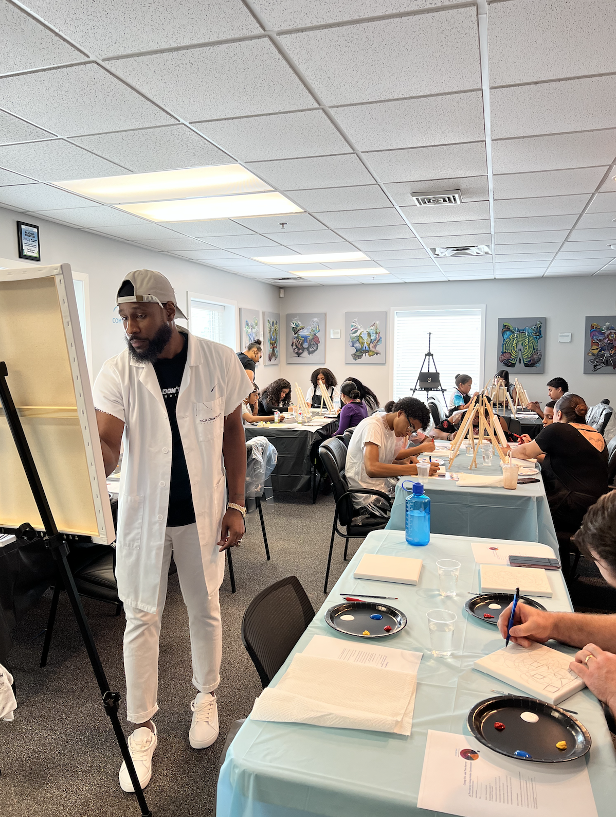 An art class with students seated at tables, painting on canvases with easels, led by an instructor. The room has artwork on the walls and a window letting in natural light.