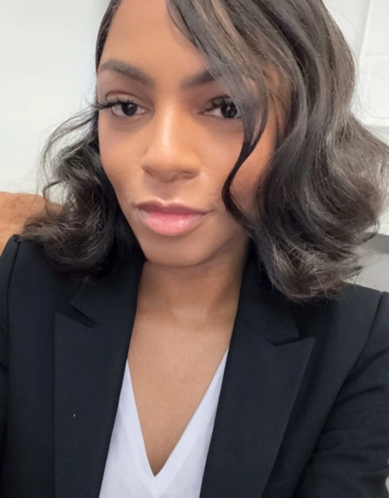 A close-up photo of a woman with medium-length wavy black hair, wearing a black blazer over a white top.