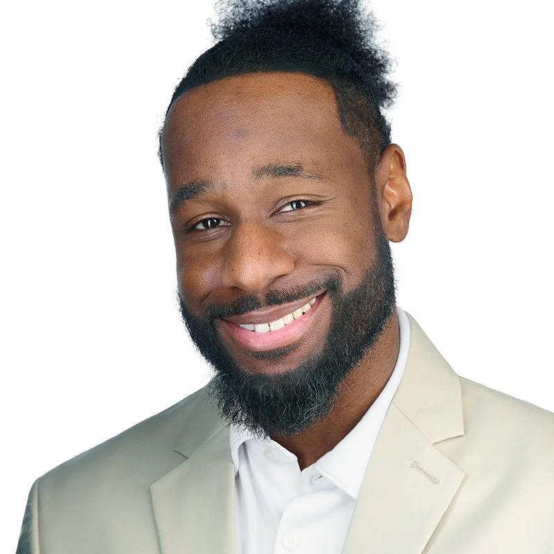 Portrait of a smiling Black man with a beard, wearing a light beige blazer and white shirt, against a white background.
