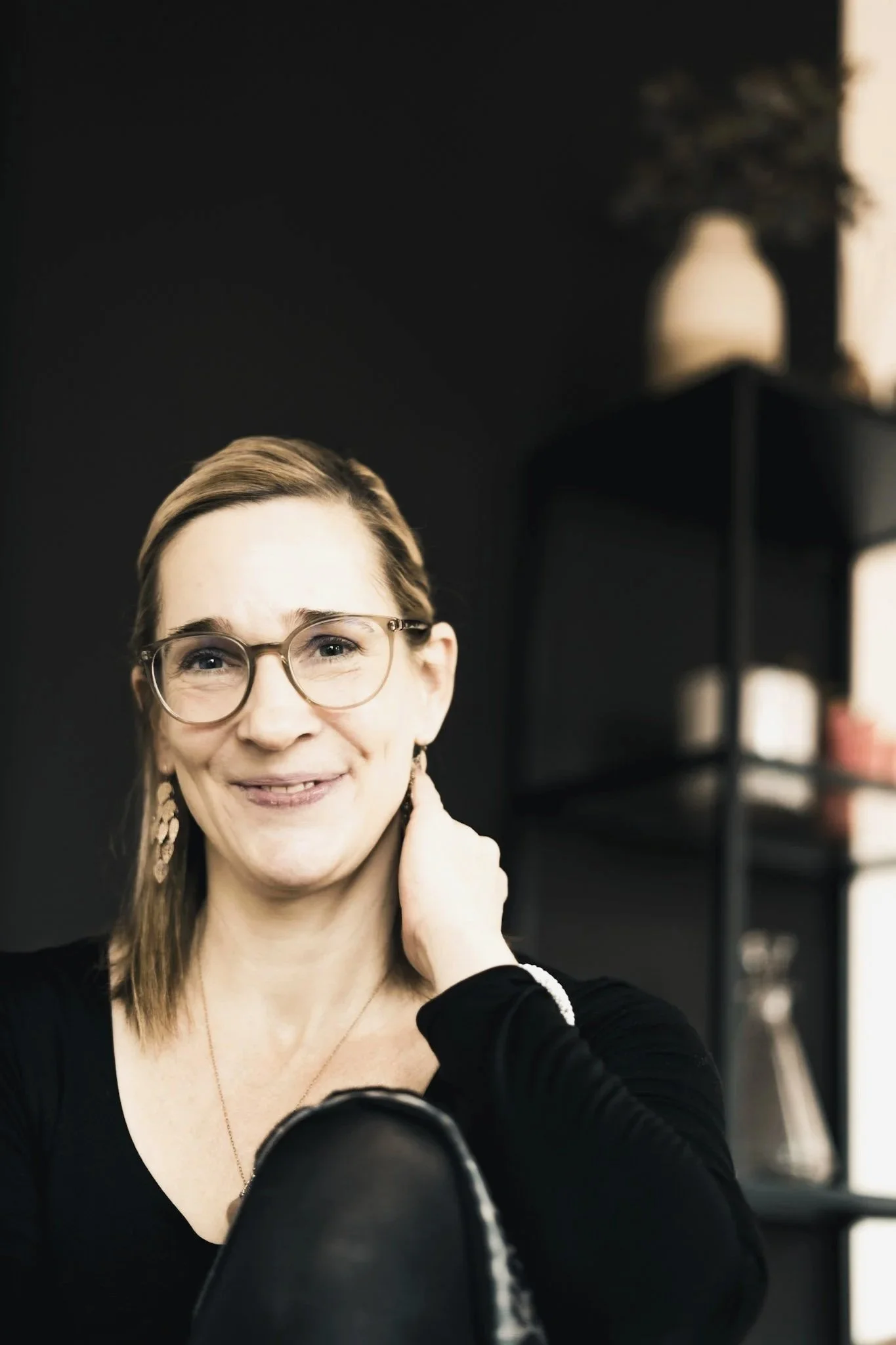 Nahaufnahme einer lächelnden Frau mit Brille, schwarzen Oberteil, im Vordergrund eine schwarze Tasche, im Hintergrund eine schwarze Regalwand mit Dekorationen, dunkler Raum mit natürlichem Licht