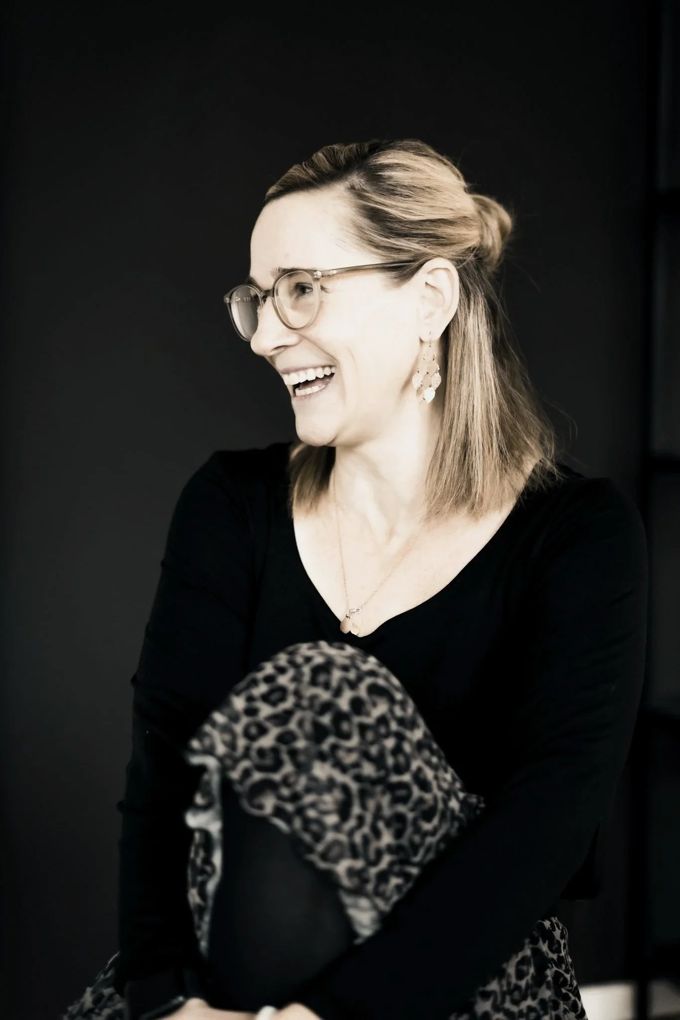 Lächelnde Frau mit Brille und Tiermuster-Hose, im schwarzen Kleid, gegen dunklen Hintergrund