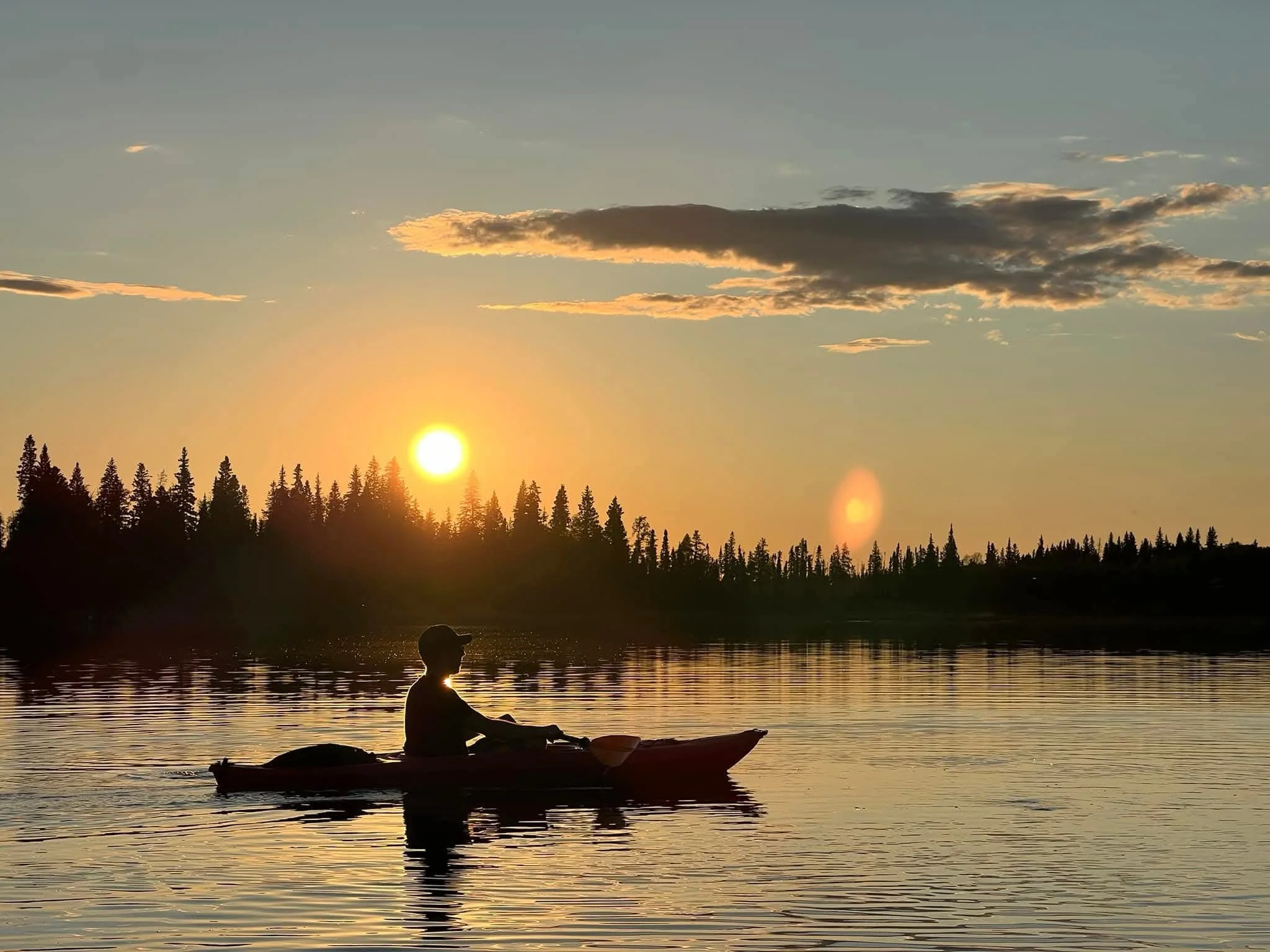 A person kayaking on calm water during sunset with a silhouette of trees and a few clouds in the sky.