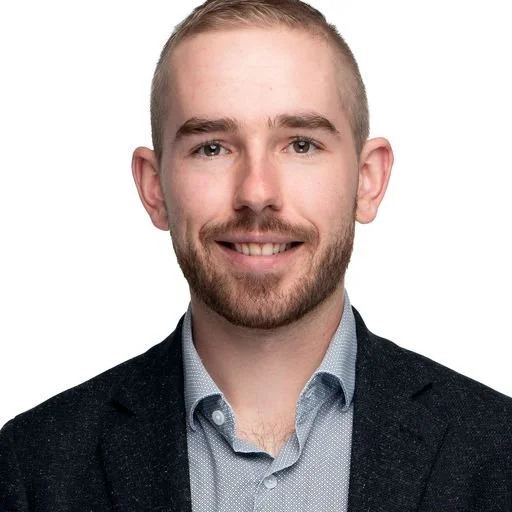 A young man with short blonde hair, a beard, and blue eyes, smiling and wearing a dark blazer over a light blue button-up shirt against a white background.