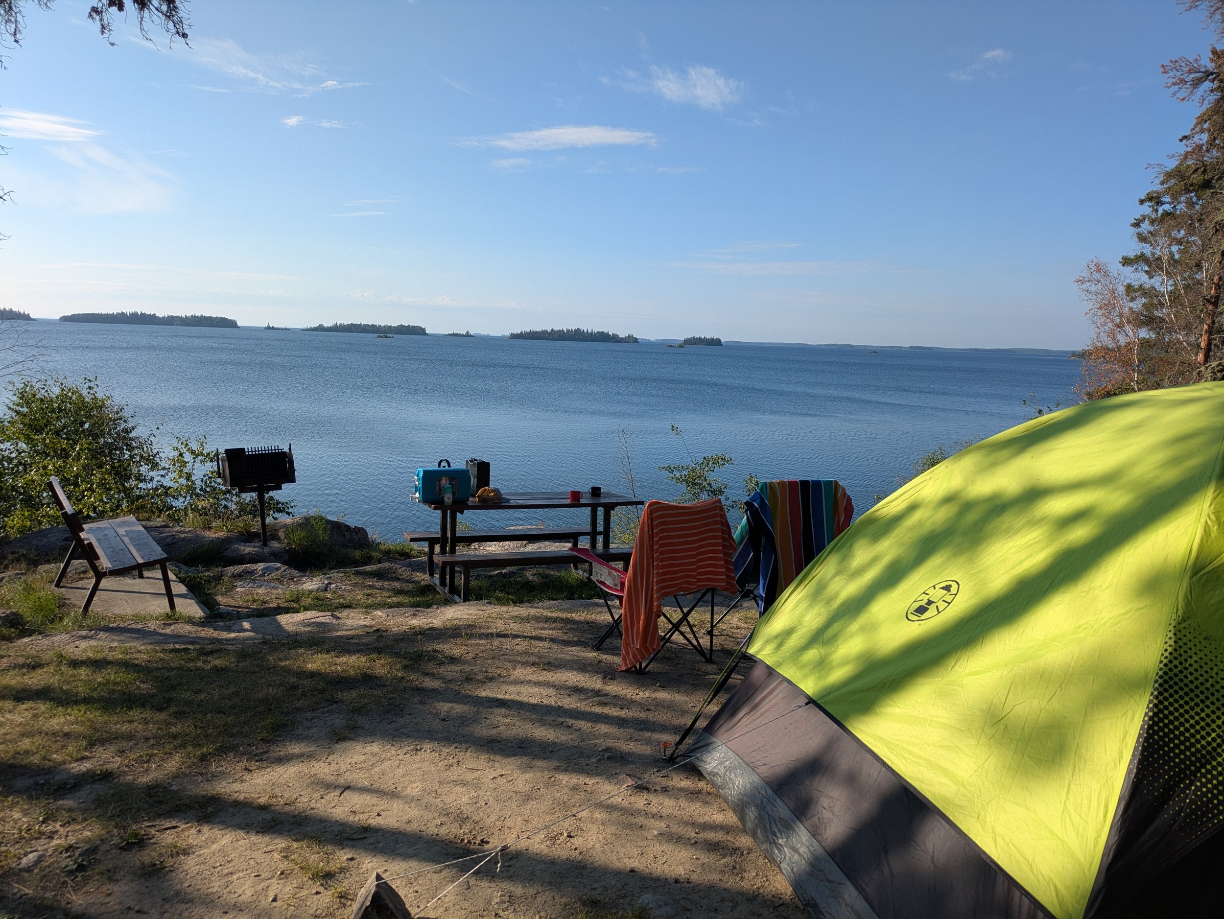 A scenic lakeside camping scene with a yellow tent, folding chairs, a picnic table, a small grill, and a view of a calm lake with islands on the horizon under a partly cloudy sky.