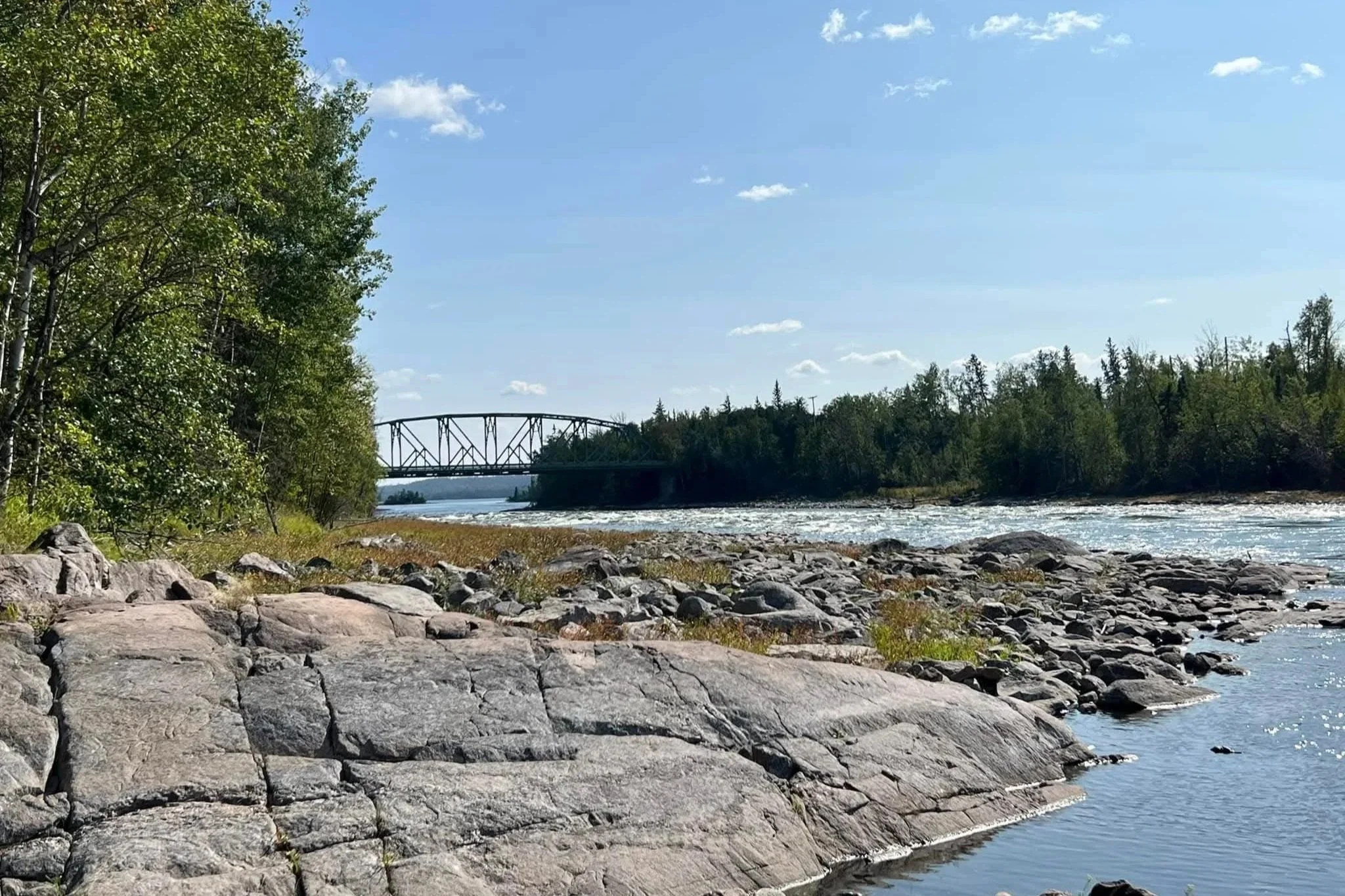 A river with rocks along the shoreline, trees on the left, a bridge in the background, and a blue sky with a few clouds.