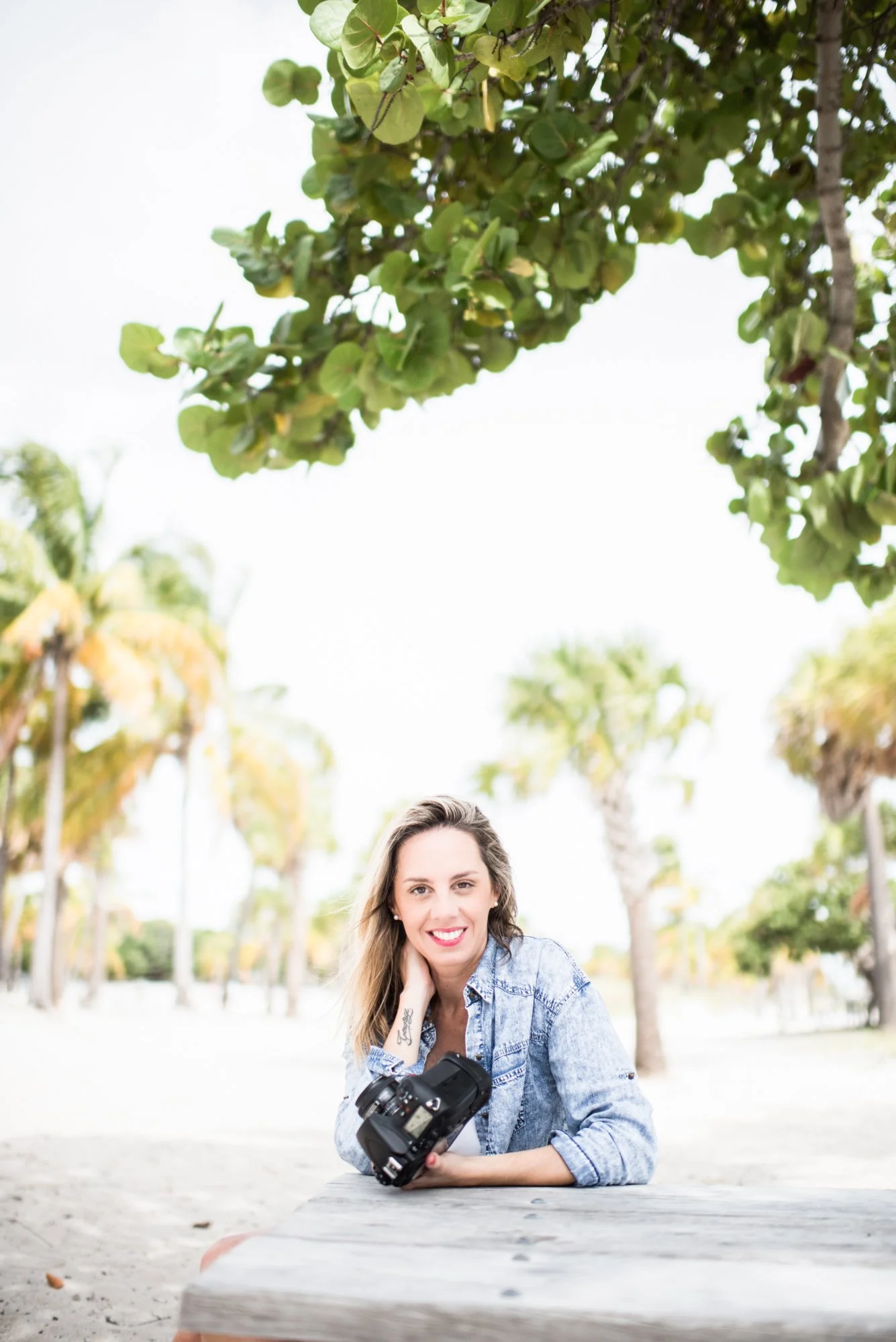 A woman with long, wavy hair, wearing a denim shirt, smiling and leaning on a wooden table outdoors under a tree. She has a camera in her hand and is looking at the camera. The background includes palm trees and a bright, clear sky.