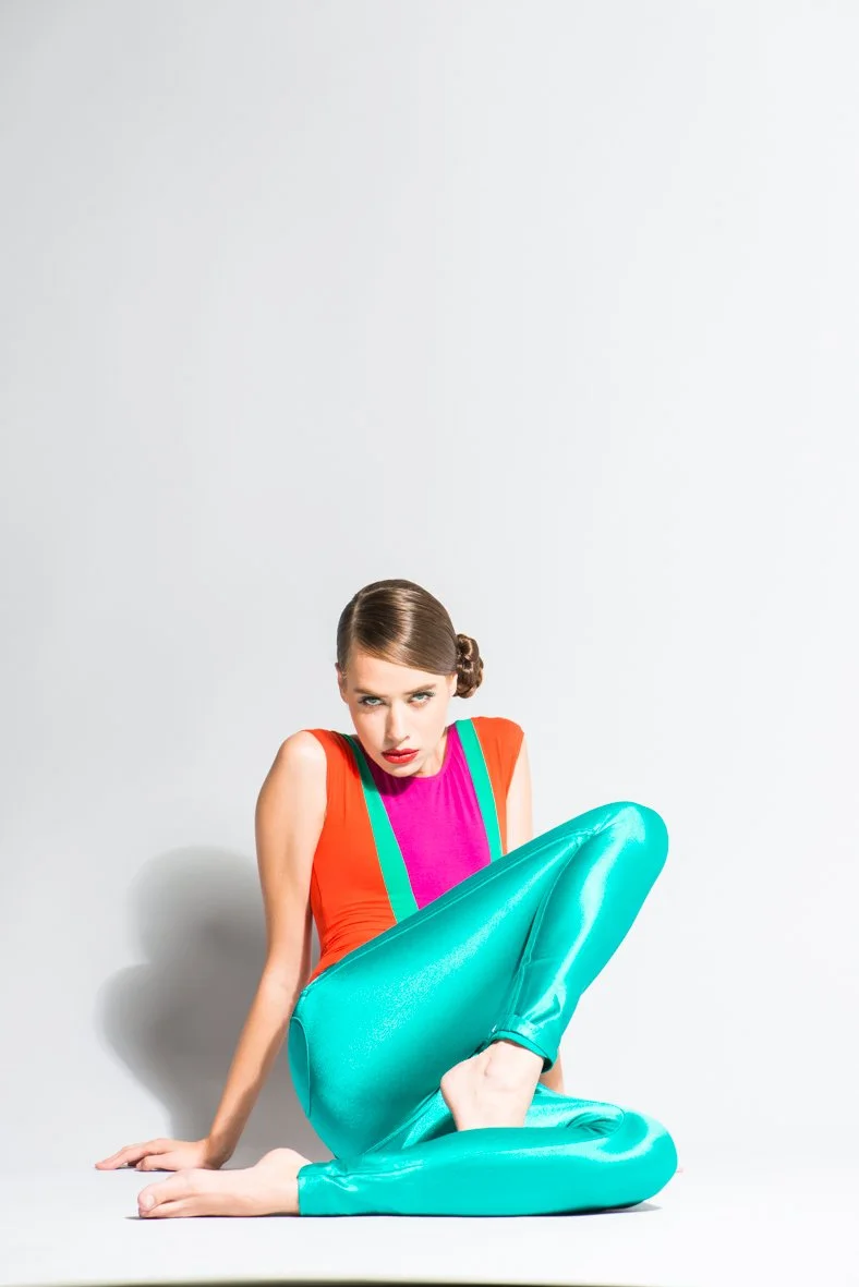 A young woman with brown hair styled in an elegant side bun, wearing a colorful sleeveless top with orange, pink, and green accents, and shiny teal pants. She is sitting on the floor with one leg bent and the other extended, looking directly at the camera with a serious expression against a plain white background.