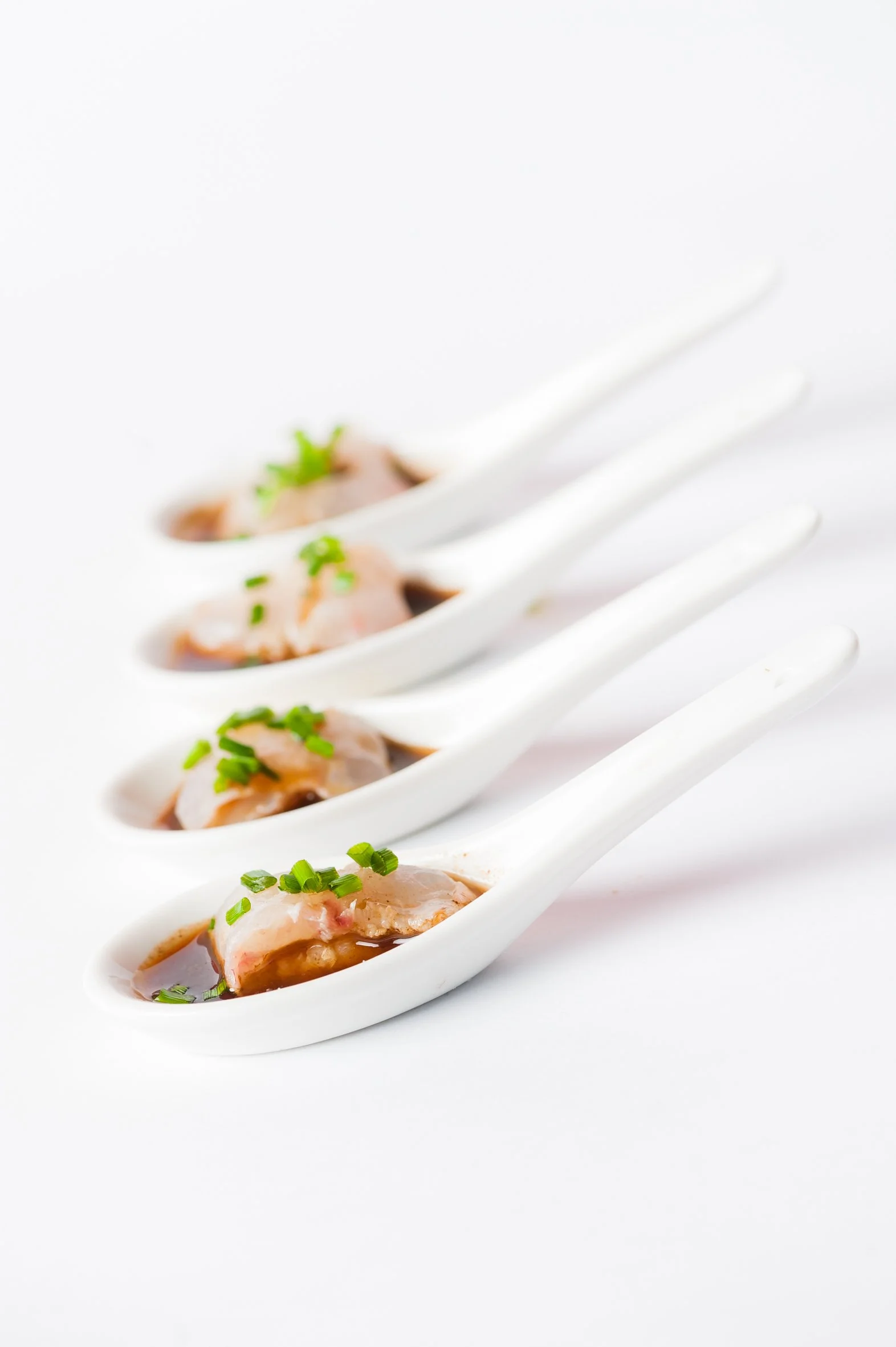 Four white ceramic spoons each containing raw fish with green herbs and brown sauce, arranged in a diagonal line on a white surface.