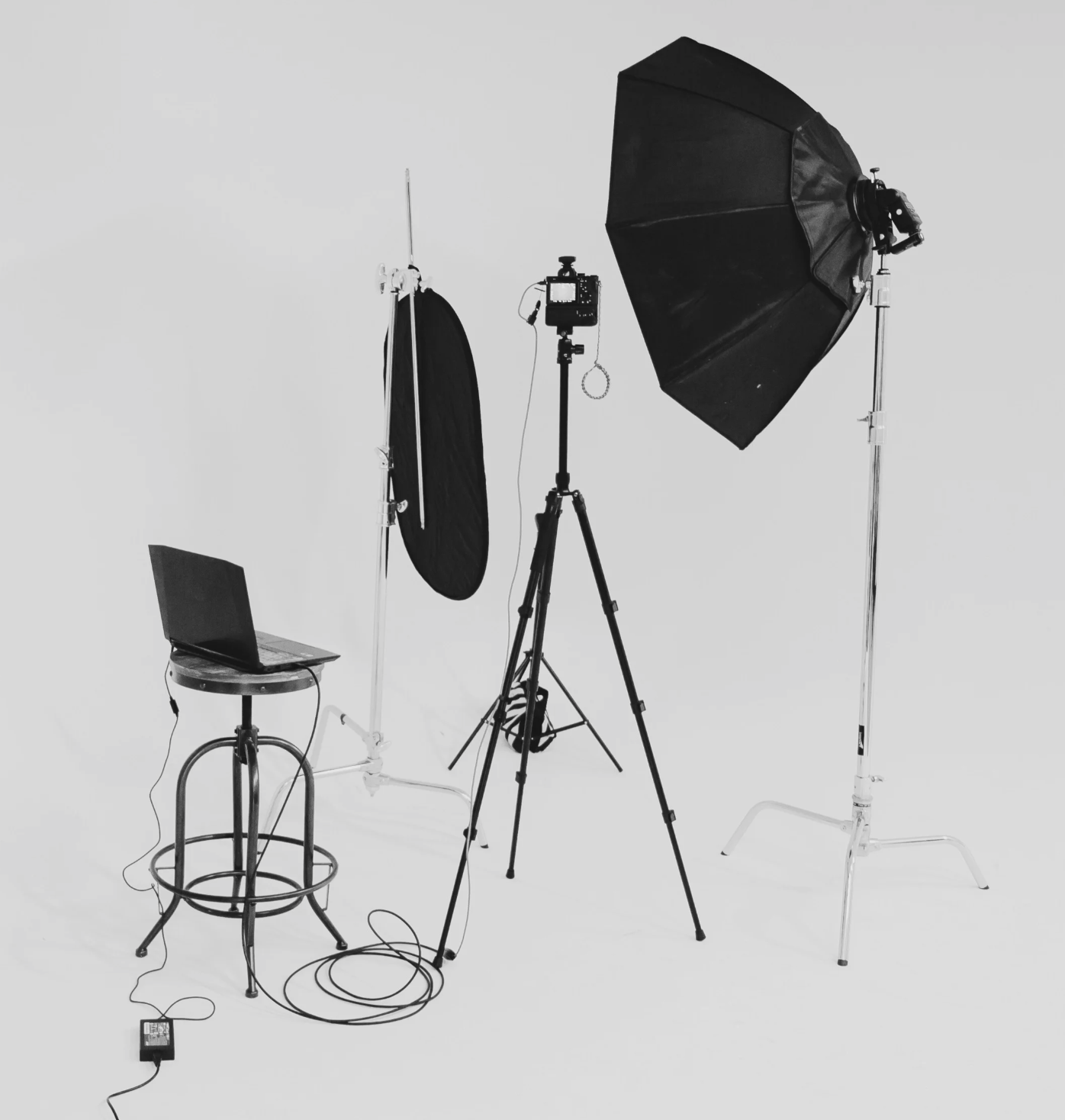 Photography studio setup with lighting equipment, a camera on a tripod, a laptop on a stool, and a white backdrop.