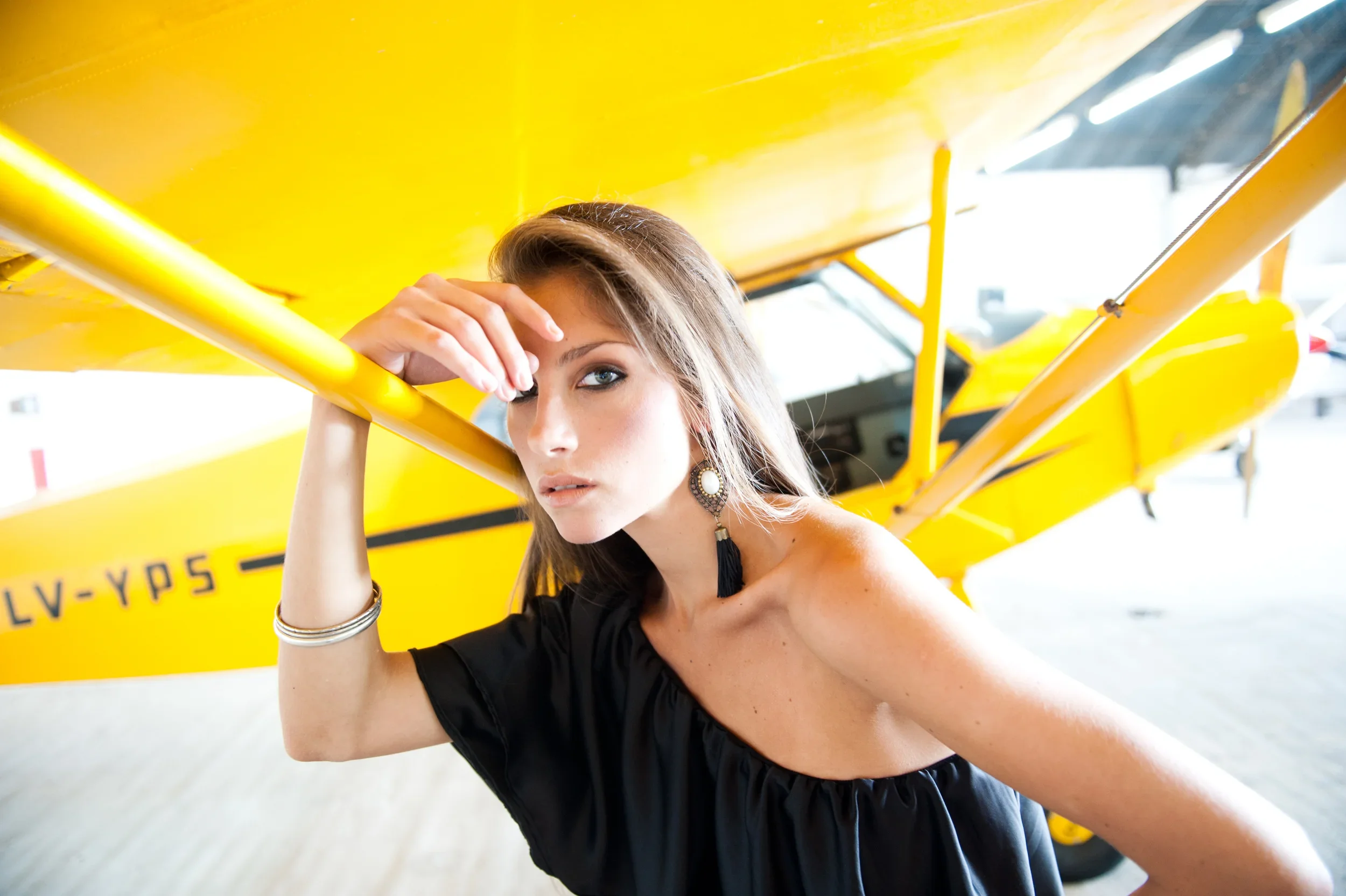 A woman with long brown hair wearing a black off-shoulder top, large earrings, and silver bracelets is posing in front of a bright yellow airplane, resting her head on her hand, and looking at the camera.