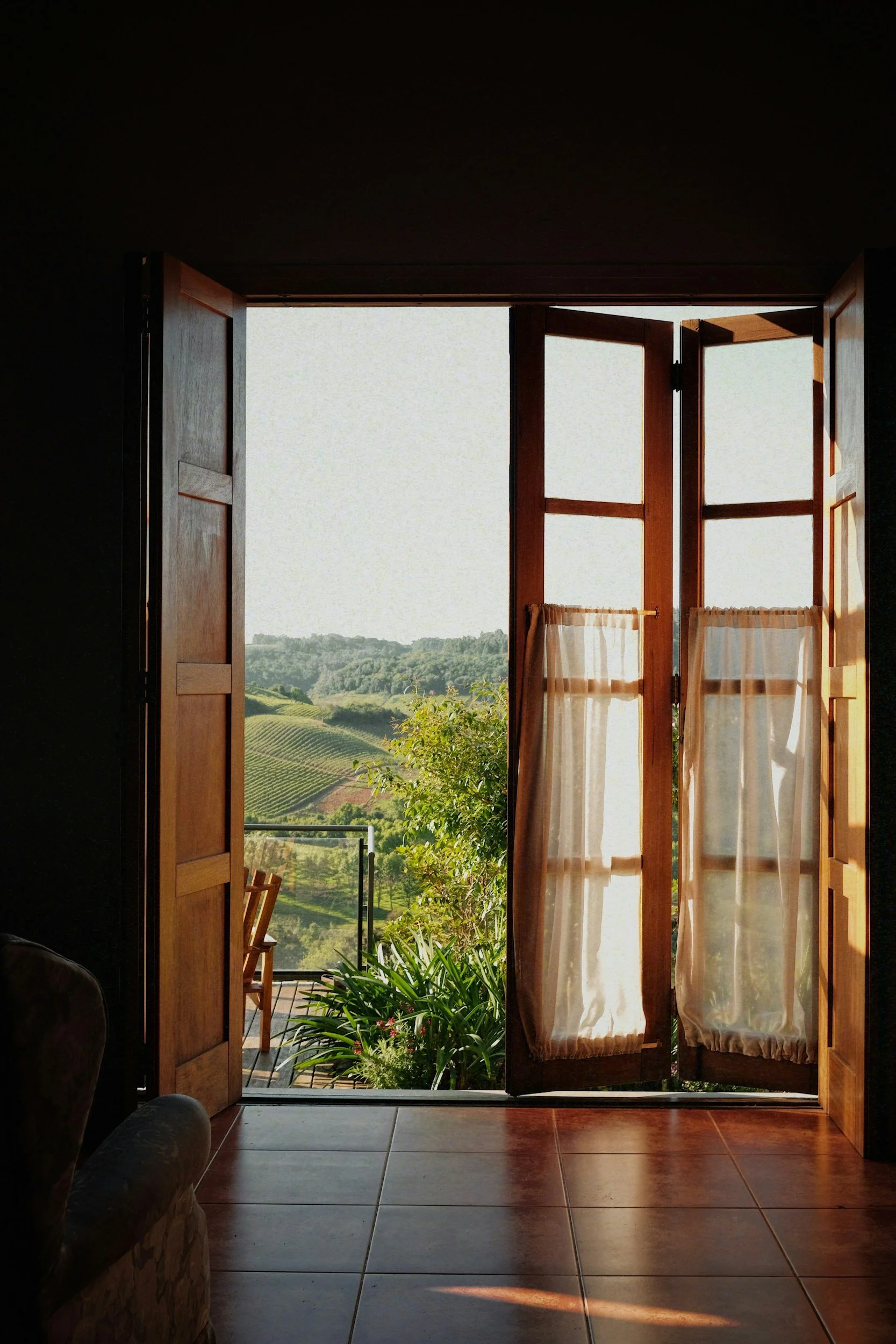 Open wooden doors leading to a lush green landscape with trees and rolling hills, viewed from inside a room with tiled floor and beige curtains.