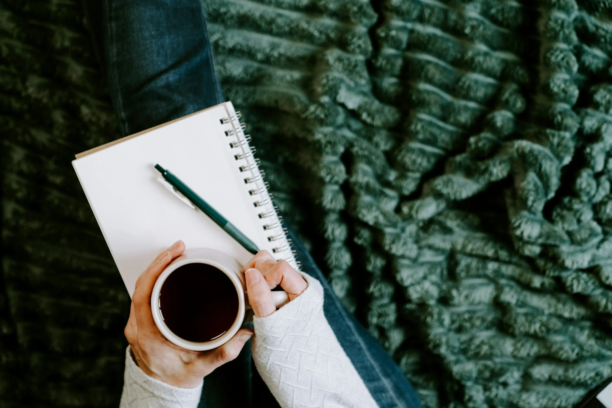 Person holding a cup of coffee and sitting outdoors, with a notebook and pen on their lap, and a fuzzy green blanket or rug underneath.