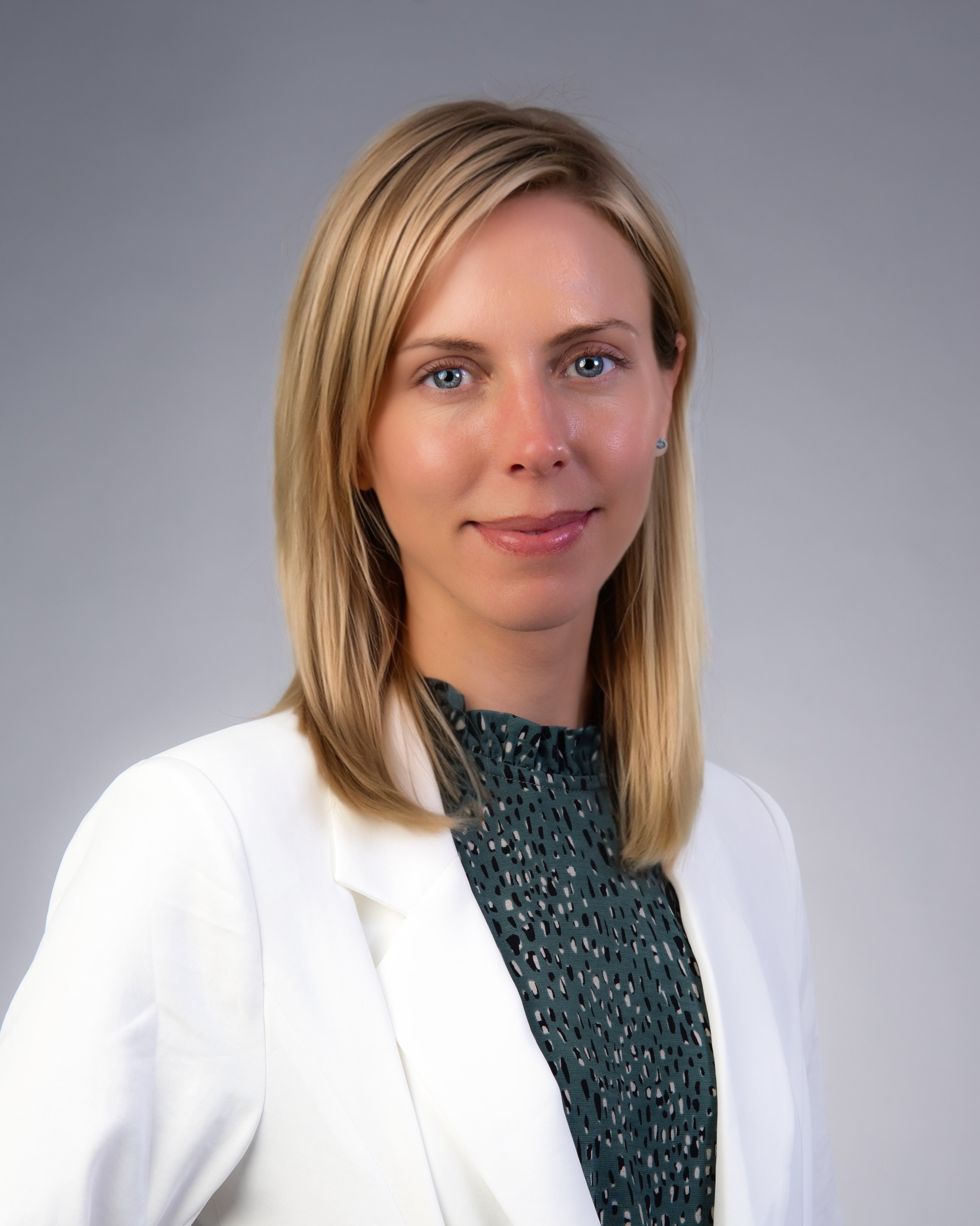 A woman with blonde hair, blue eyes, and light skin, wearing a white blazer over a dark patterned blouse, standing against a neutral background.