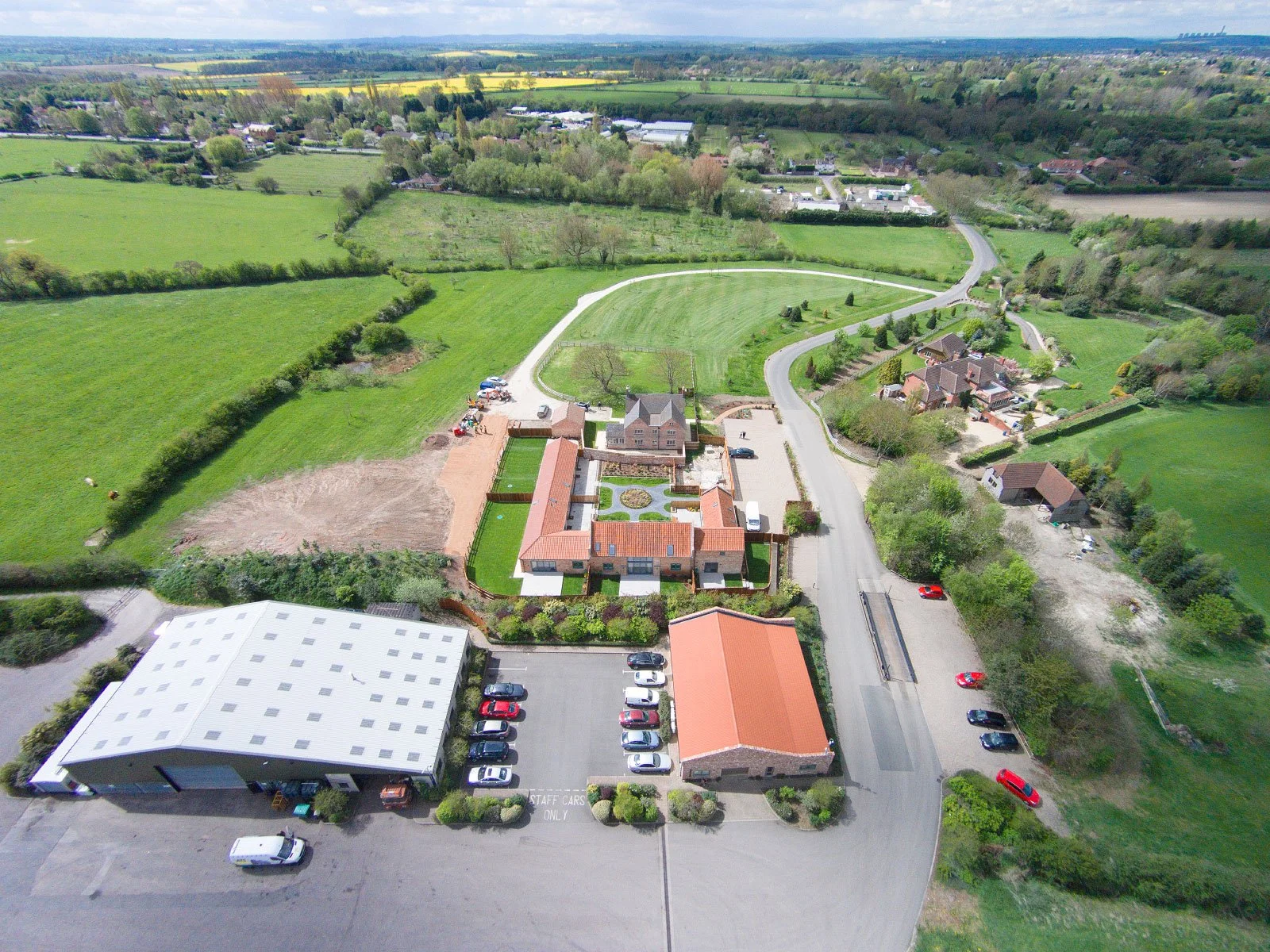 Aerial view of a countryside estate with a large red-roofed house, surrounding greenery, a parking lot, and open fields.