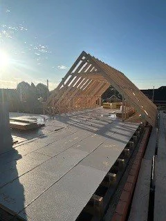 Construction site with a wooden roof frame under construction, clear blue sky, and sunlight reflecting off the building materials.