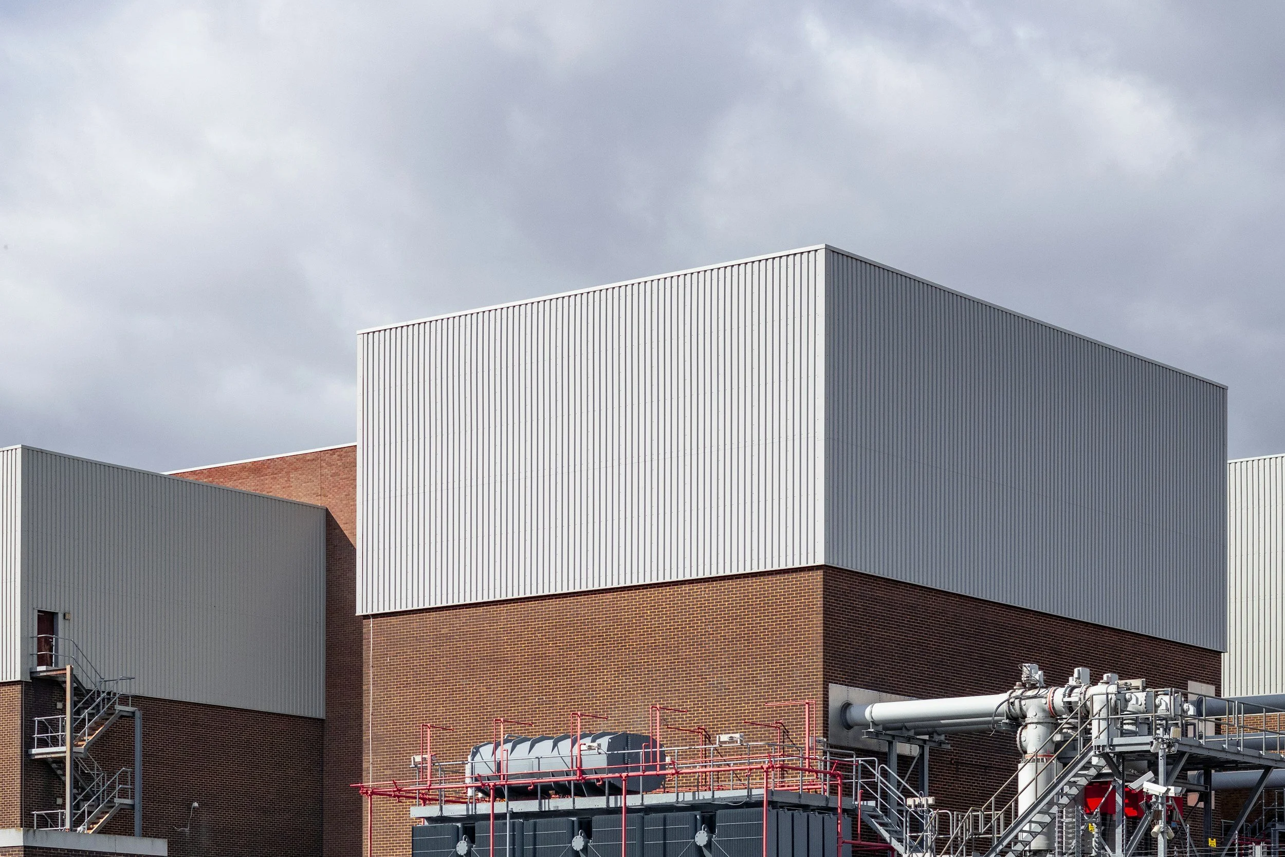 Industrial building with metal siding and brick section, industrial pipes and machinery outside, under cloudy sky.