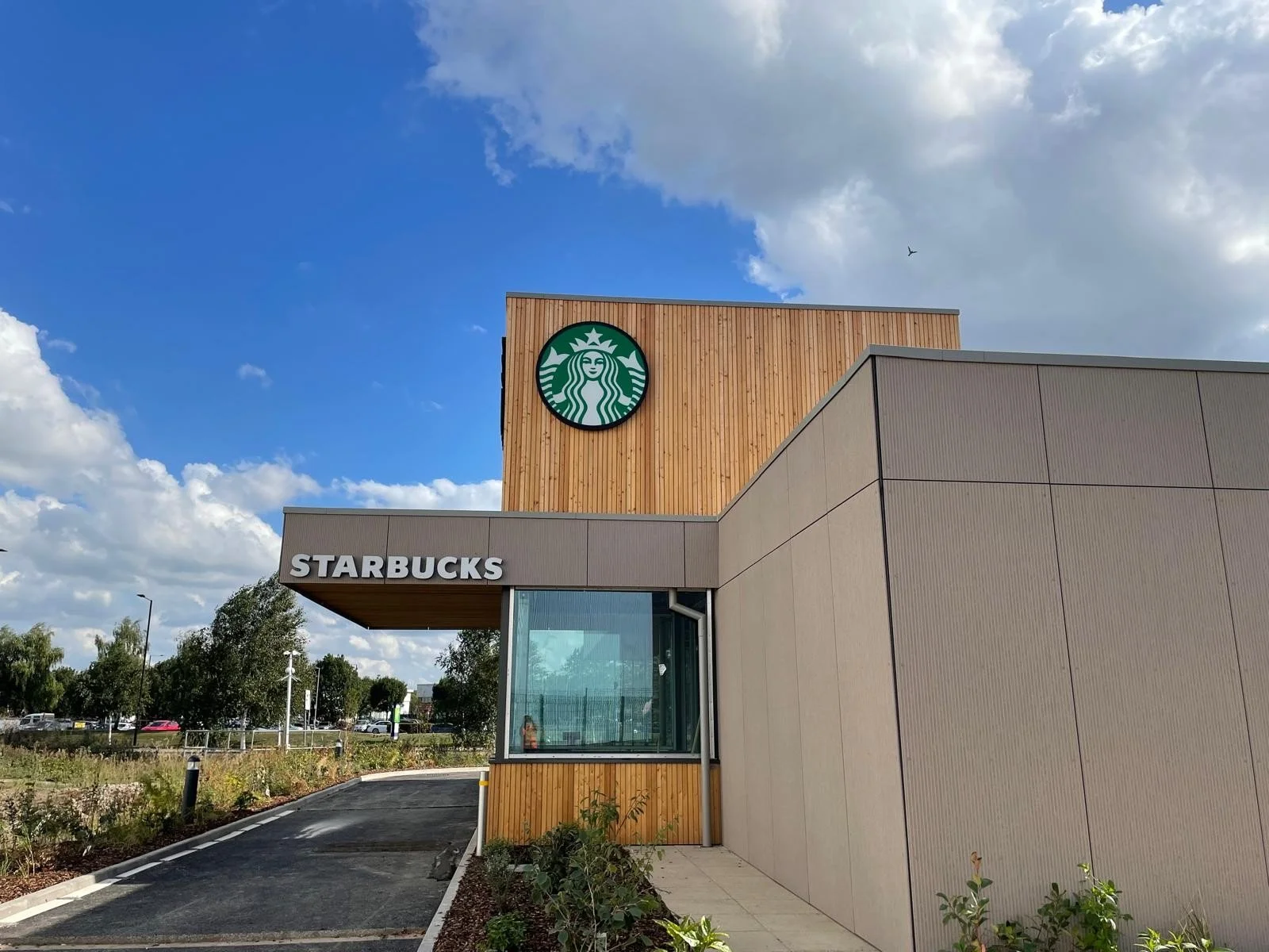 Exterior view of a Starbucks store with wood and beige panels, a large green Starbucks logo, and a sign with white letters spelling 'STARBUCKS' under a partly cloudy sky.