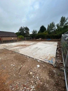 Empty construction site with a concrete slab, surrounded by dirt and a metal fence, with trees and houses in the background under a cloudy sky.