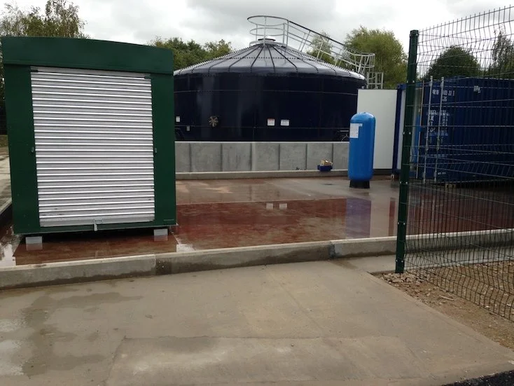 Industrial water storage tank with a metal staircase, a blue compressed air tank, and a fenced area with a roller shutter container.