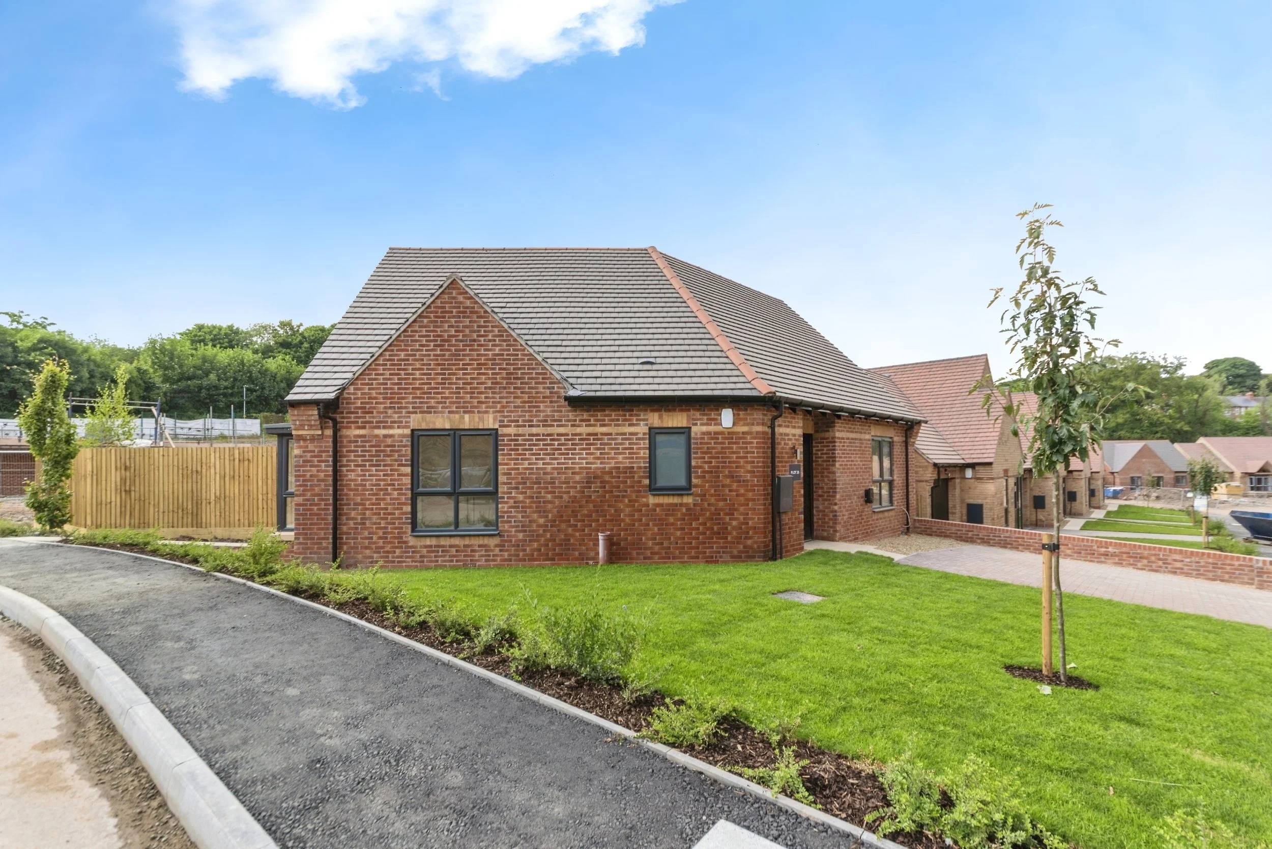 Newly constructed brick house with gray tiled roof, black window frames, a small front lawn with a young tree, and a paved pathway in a suburban neighborhood.
