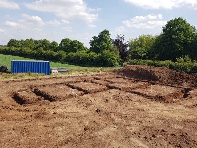 Construction site with dug-out rectangular areas in dirt, a large mound of soil, trees in the background, and a blue shipping container on the left side.