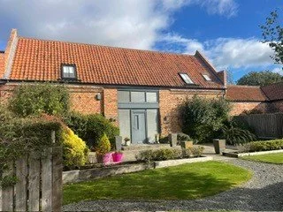 Brick house with orange tiled roof, front garden with grass and potted plants, under blue sky with clouds.