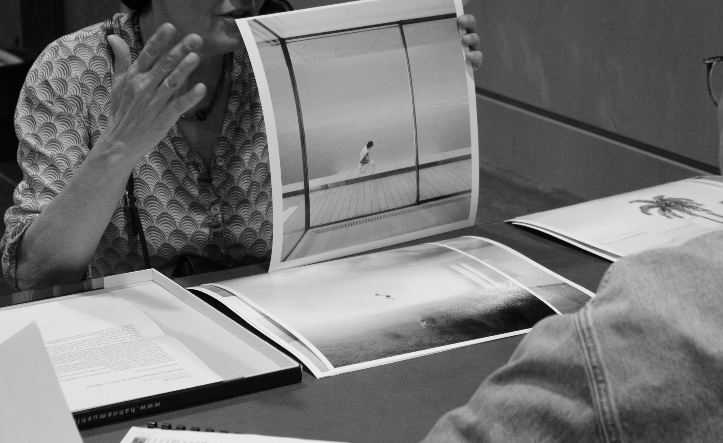 Une femme montre un album photo noir et blanc où l'on voit une personne seule dans un espace ouvert, probablement une plage ou un parc. D'autres pages de livres ou magazines sont également sur la table.