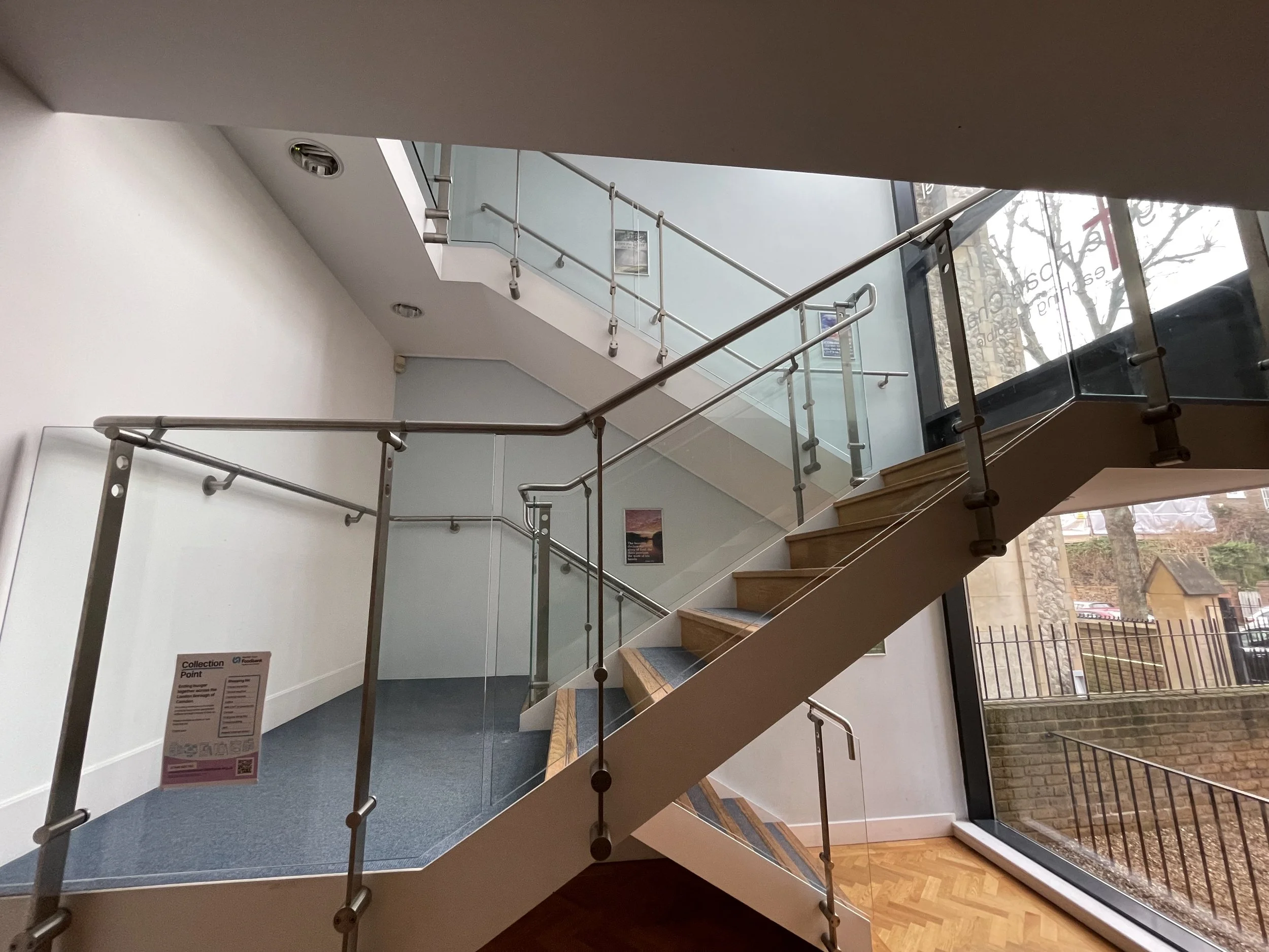 Interior of a modern building with a staircase featuring wooden steps, glass railings, and metal handrails. Large windows reveal an outdoor brick wall and trees.