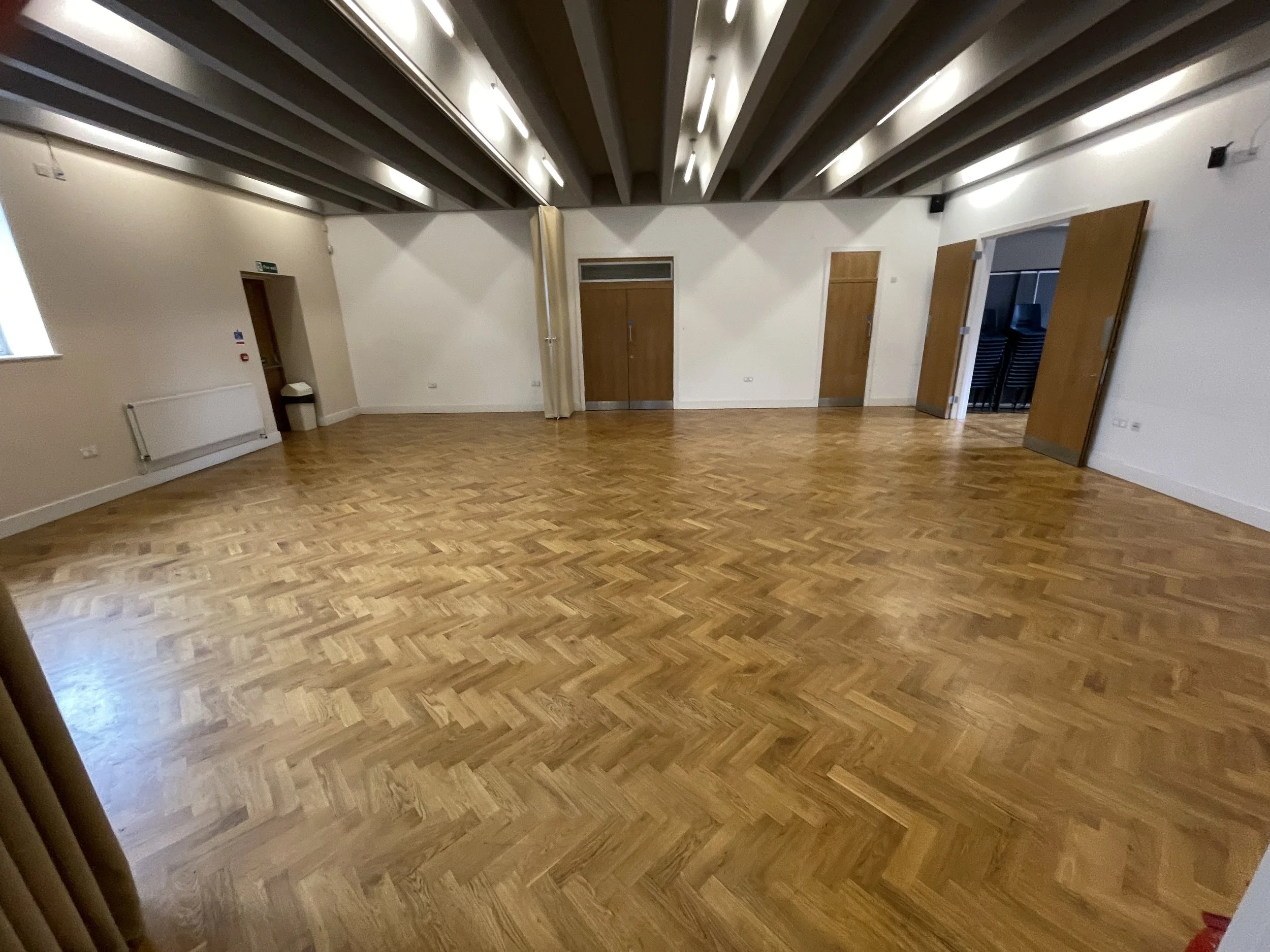 Empty room with a wooden parquet floor, white walls, and ceiling with exposed beams, with multiple doors and a window.