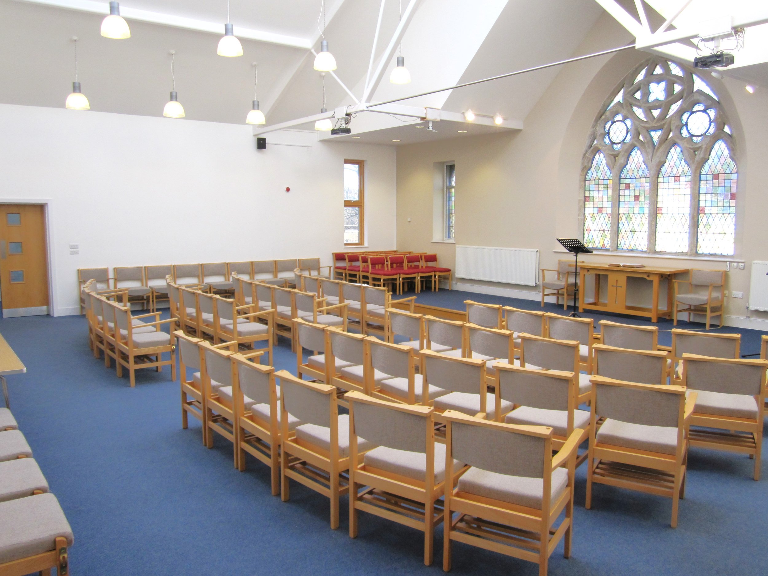 Empty church or chapel with rows of wooden chairs with gray cushions, a stage area with a music stand, large stained-glass window, and high ceiling with hanging lights.