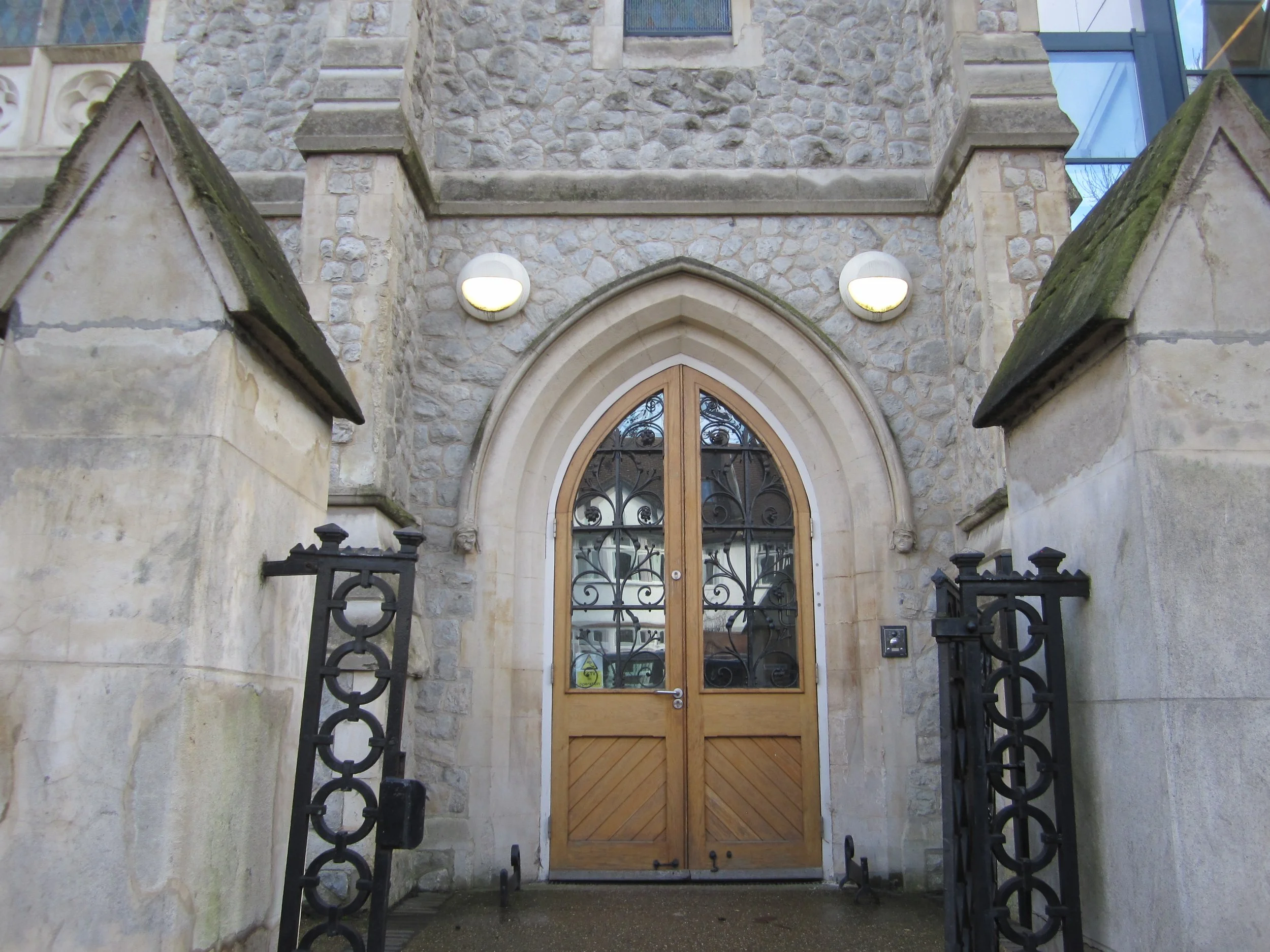 Stone and brick arched doorway with wooden double doors and ornate glass window panels, flanked by stone walls and black wrought iron gates, topped with wall-mounted lights.