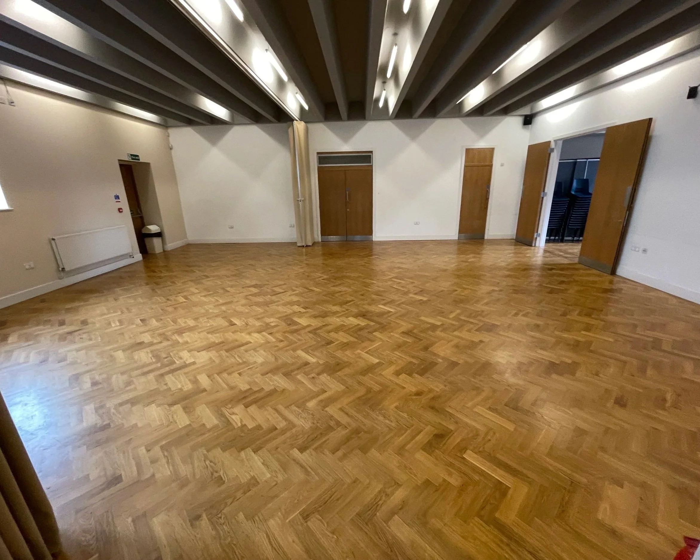 Empty room with hardwood floor, white walls, wooden doors, and ceiling beams, with some stacked chairs visible through an open door.