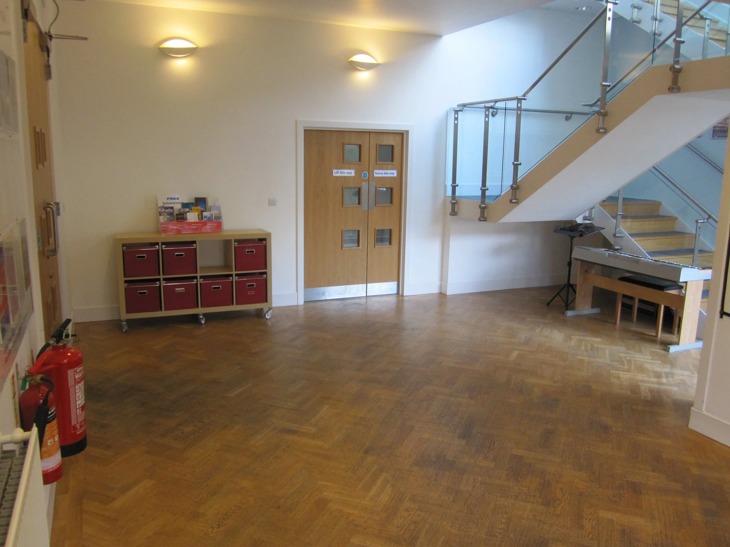 Empty classroom or lobby area with wooden flooring, a small red and wooden storage unit, a wooden door with windows, stairs with glass and metal railing, and a keyboard on a stand.