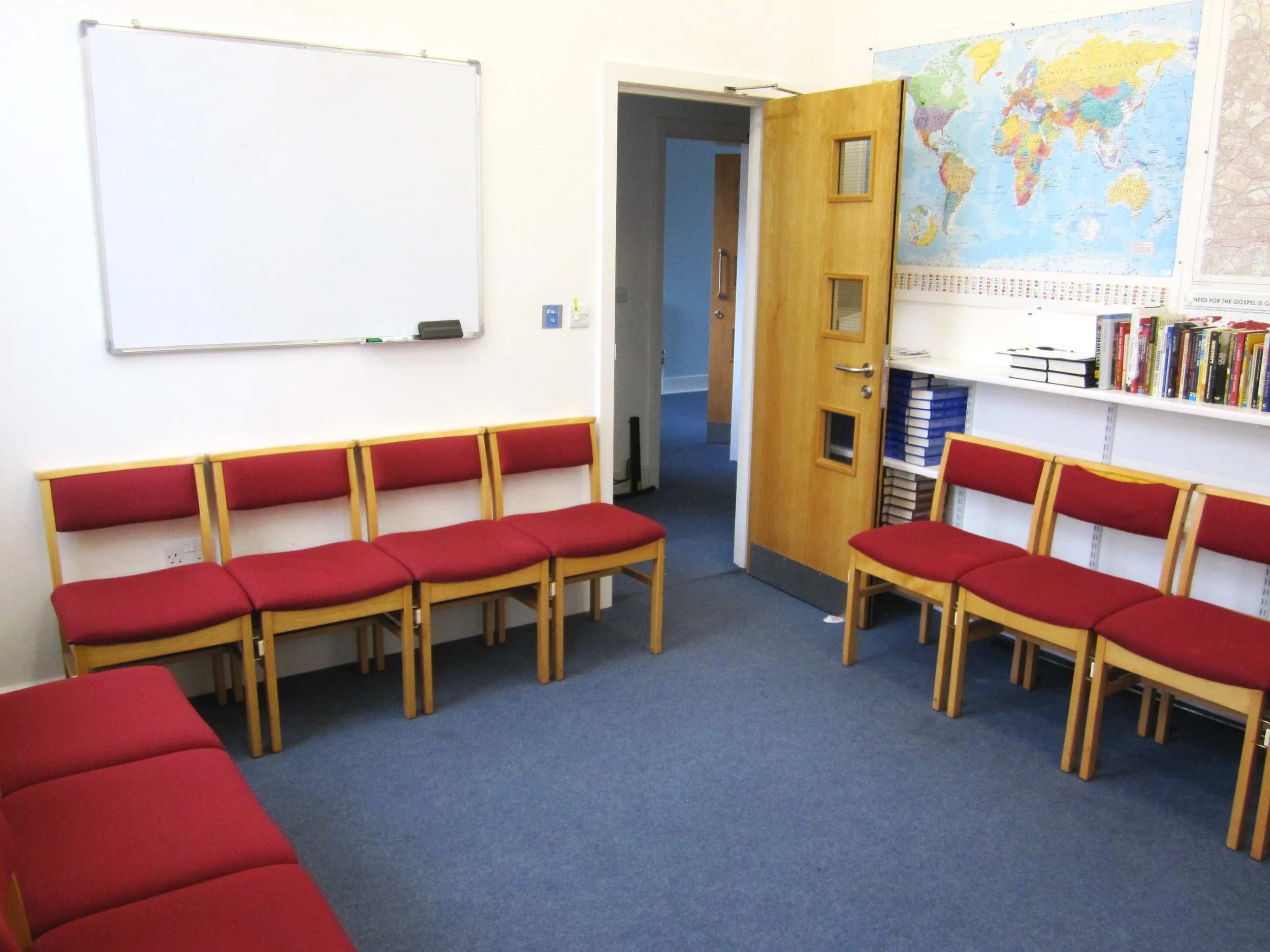 Empty classroom with red chairs arranged in a U-shape, whiteboard on the wall, open wooden door, world map, and bookshelf with books and folders.