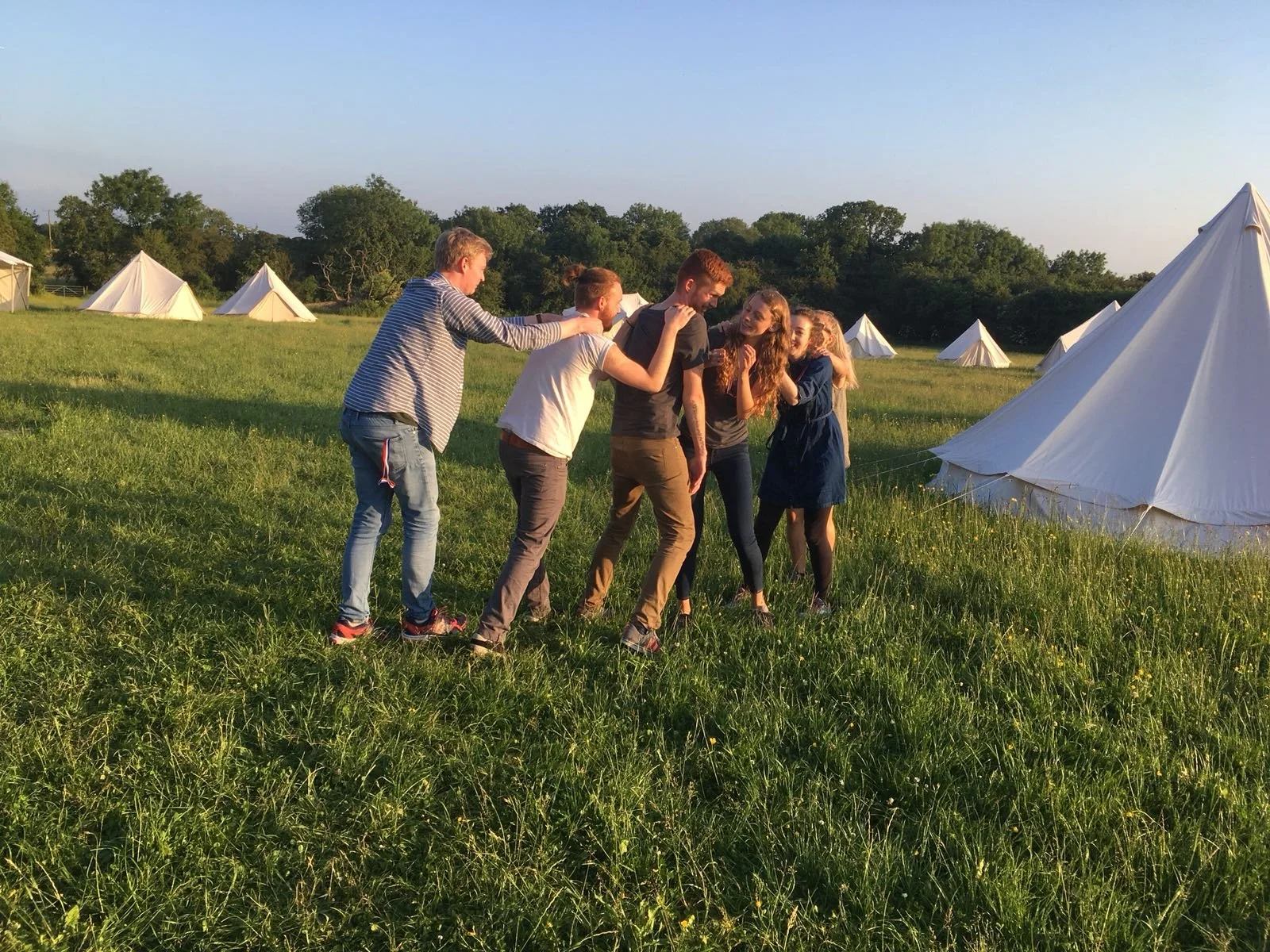 Group of six friends playing tug of war with a rope on a grassy field during sunset, with white tents in the background.