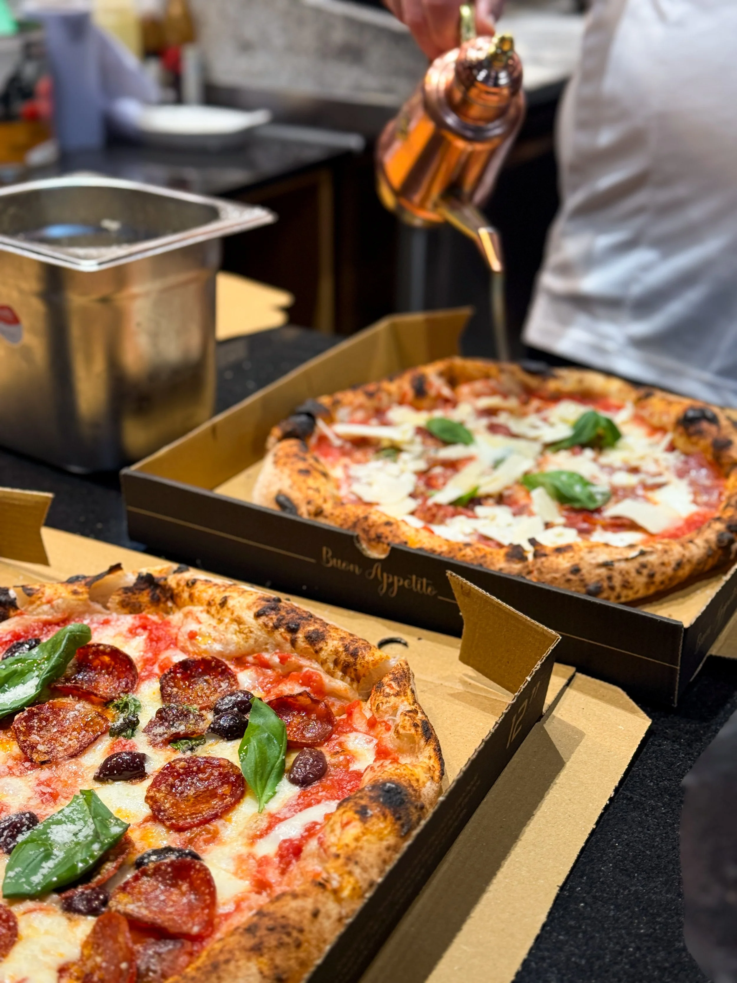Two pizzas in open boxes, one topped with pepperoni, black olives, basil, and mozzarella, the other with mozzarella, tomato sauce, parmesan shavings and basil. The Chef is pouring olive oil on the pizzas in a pizzeria setting.