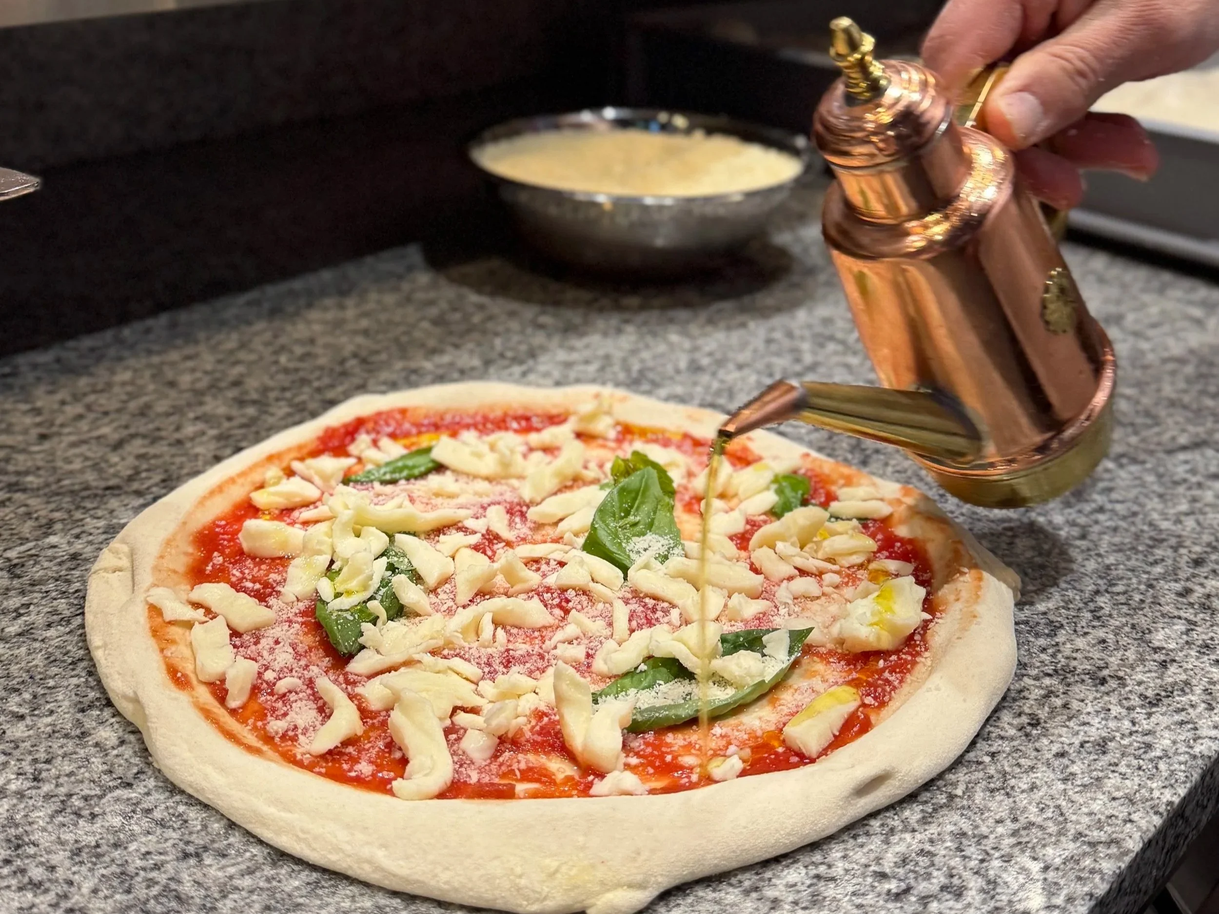 Chef pouring olive oil over a pizza topped with cheese, tomato sauce, and basil leaves.
