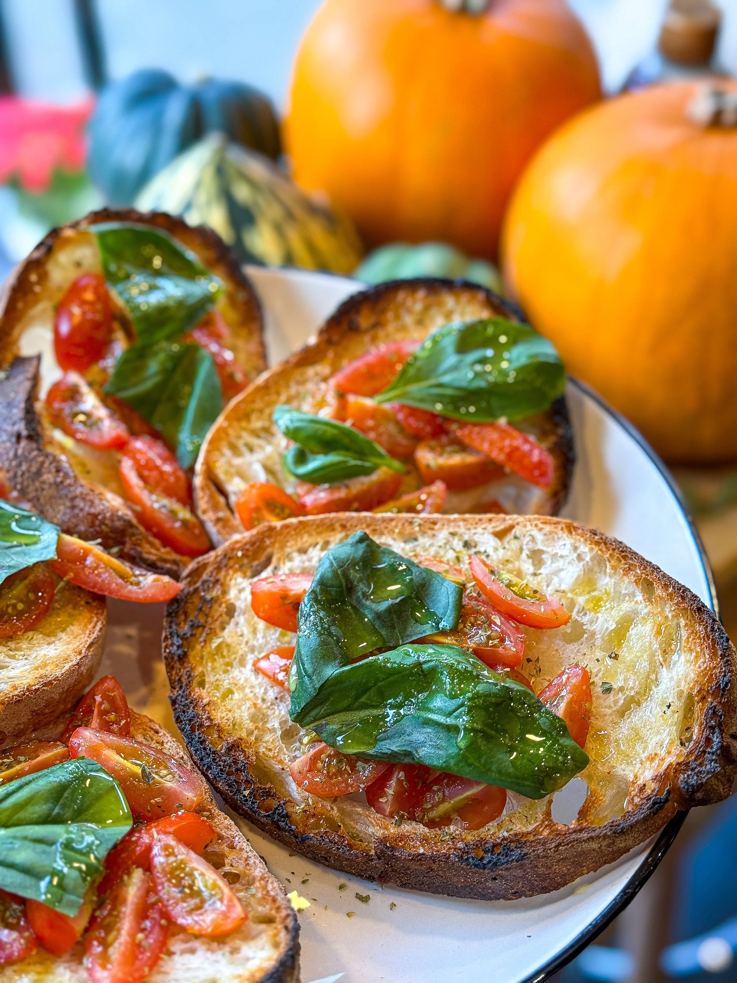 Close-up of classic Bruschettas topped with cherry tomatoes, garlic, oregano and fresh basil leaves, served on a white plate with pumpkins in the background.