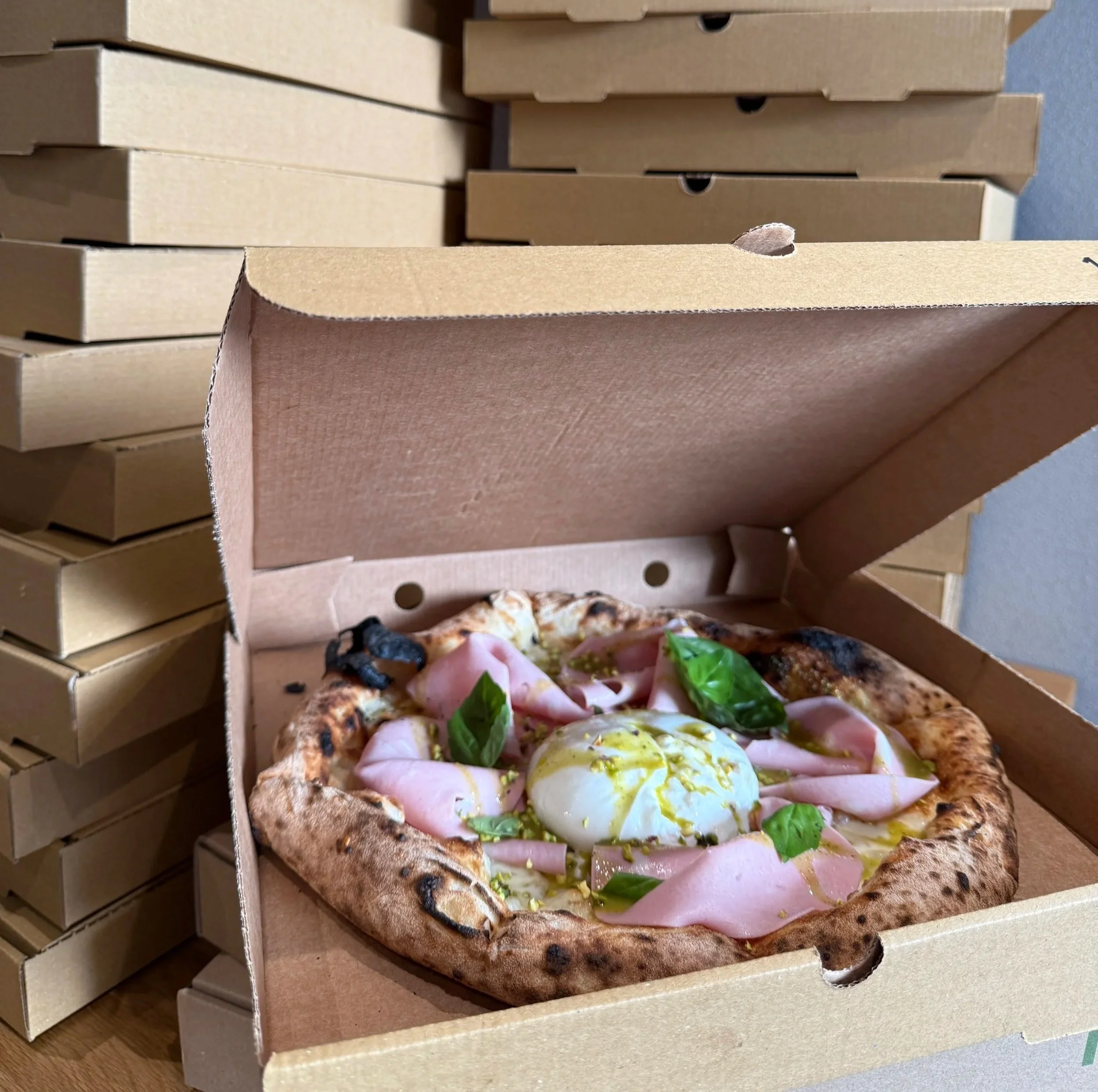 A pizza topped with mortadella, basil, pistachios pesto and chopped pistachios, and a soft-creamy burrata cheese in a cardboard pizza box, with stacked pizza boxes in the background.