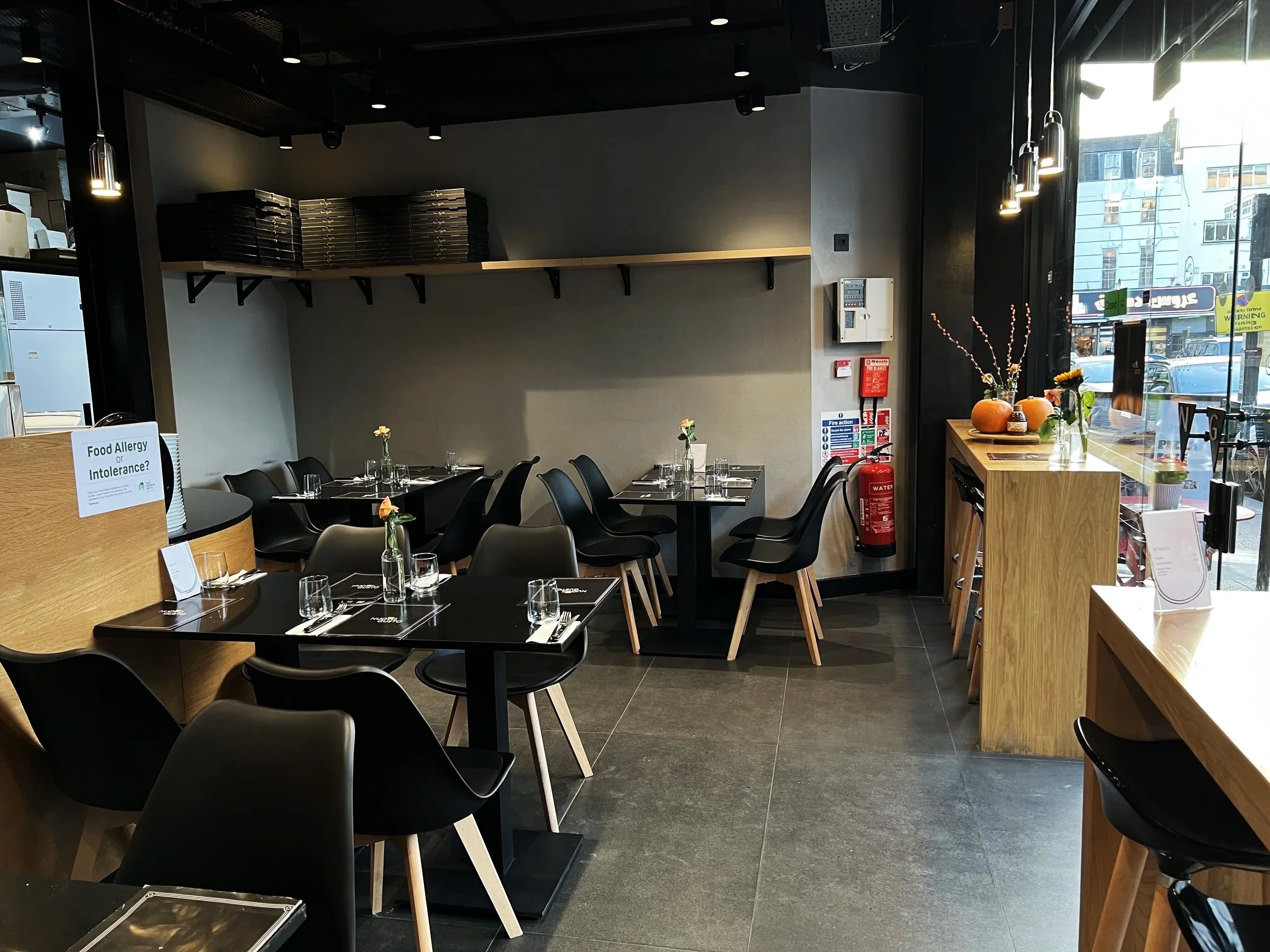 Nuovo Gusto restaurant Interiors with black tables, black and wooden chairs, and minimal decor, including flowers on tables and pumpkins on a wooden counter near the window.