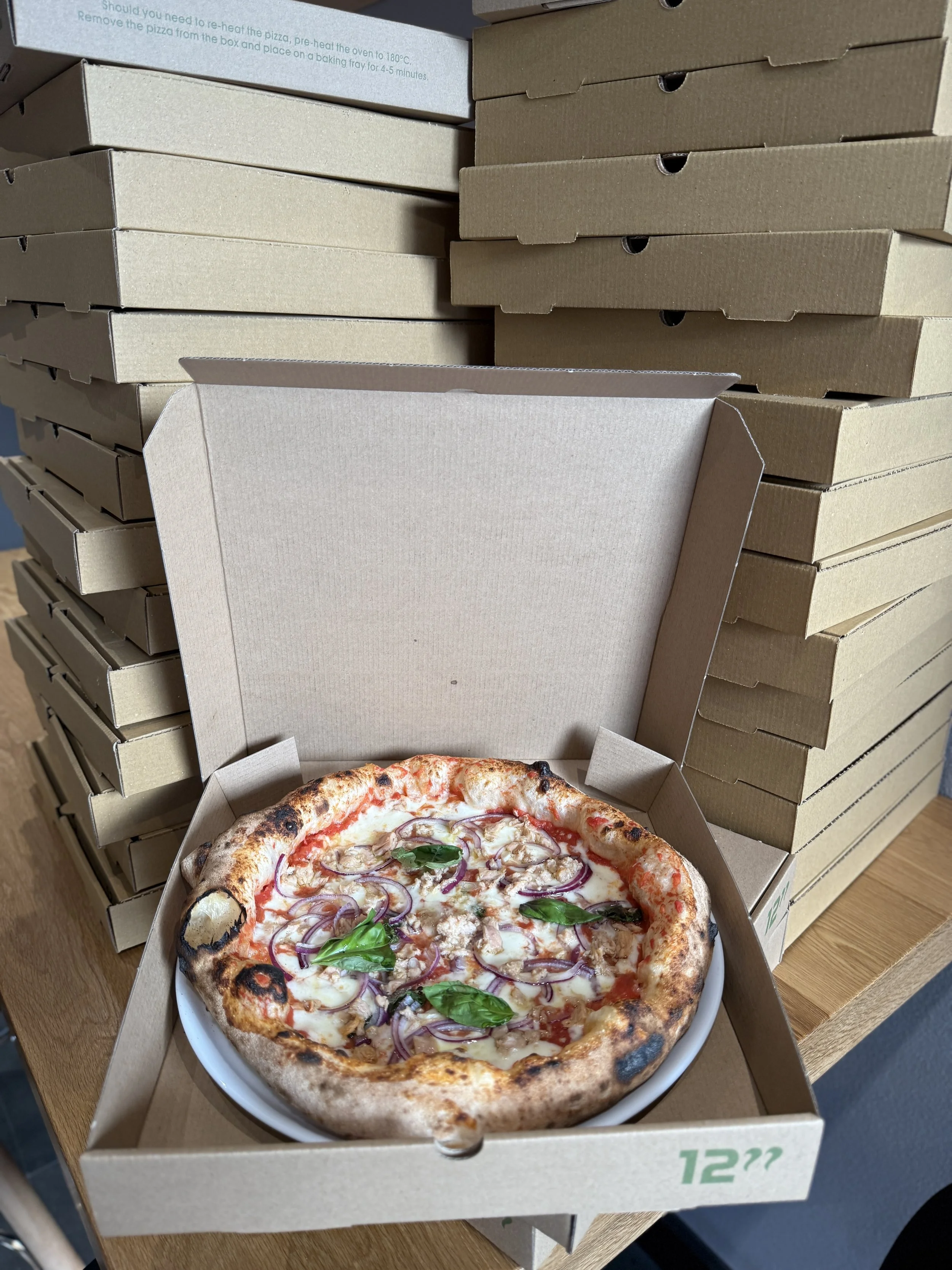 An open pizza box with a 12-inch pizza topped with mozzarella, tuna, red onions, tomato sauce, and basil leaves, placed in a pizza box surrounded by stacked empty pizza boxes.