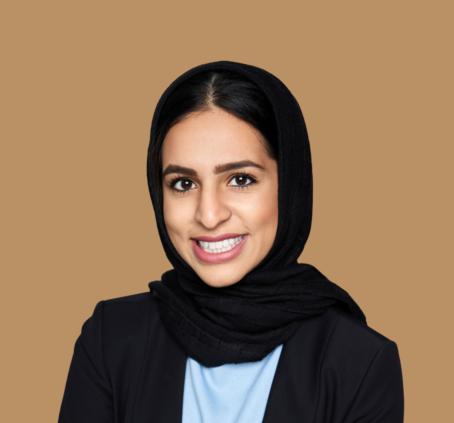 A woman with dark hair, brown eyes, and a light complexion, smiling and wearing a black headscarf, black blazer, and light blue top, against a plain beige background.