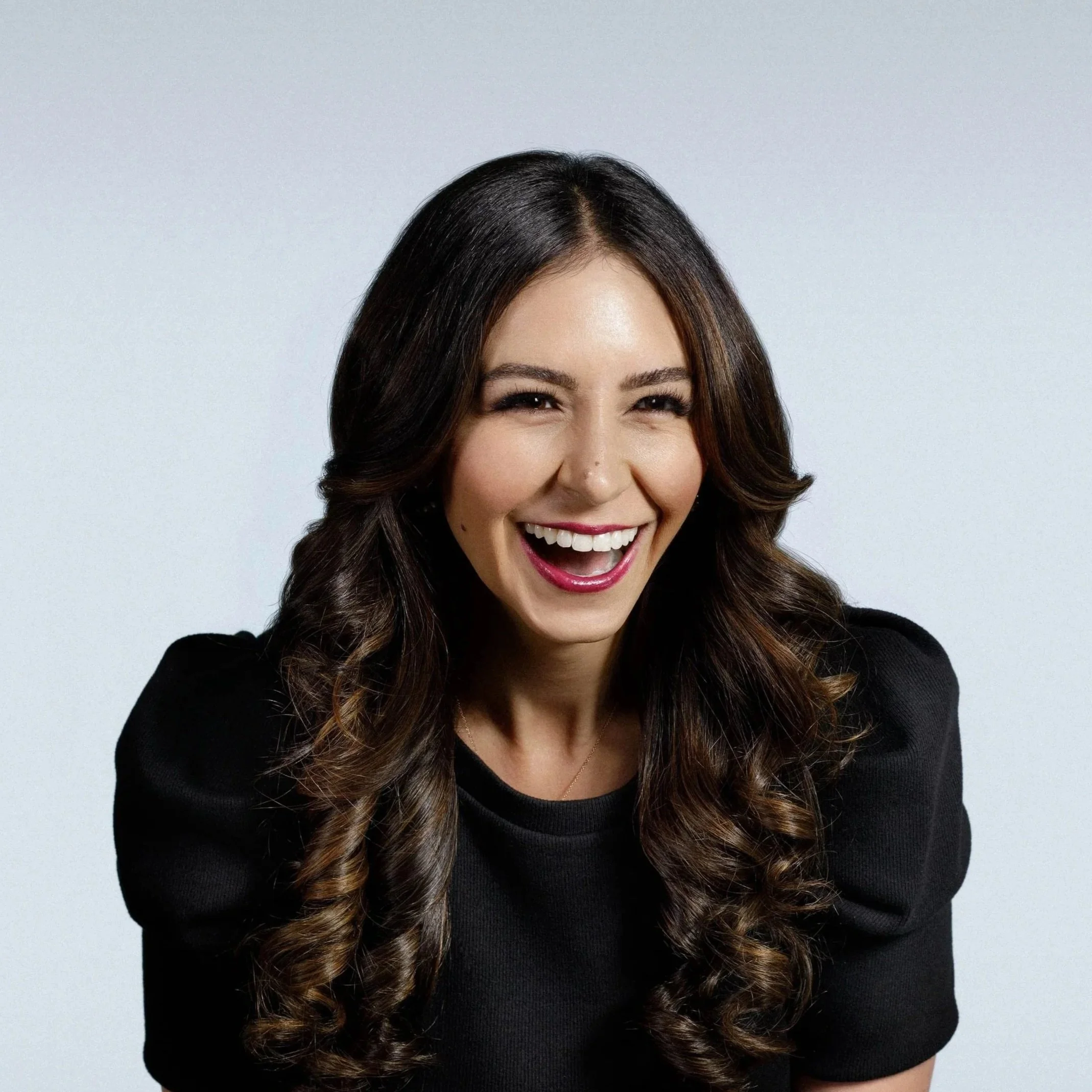 A woman with long, dark hair styled in loose curls, smiling broadly with her teeth visible, wearing a black top with puffed sleeves, against a light gray background.