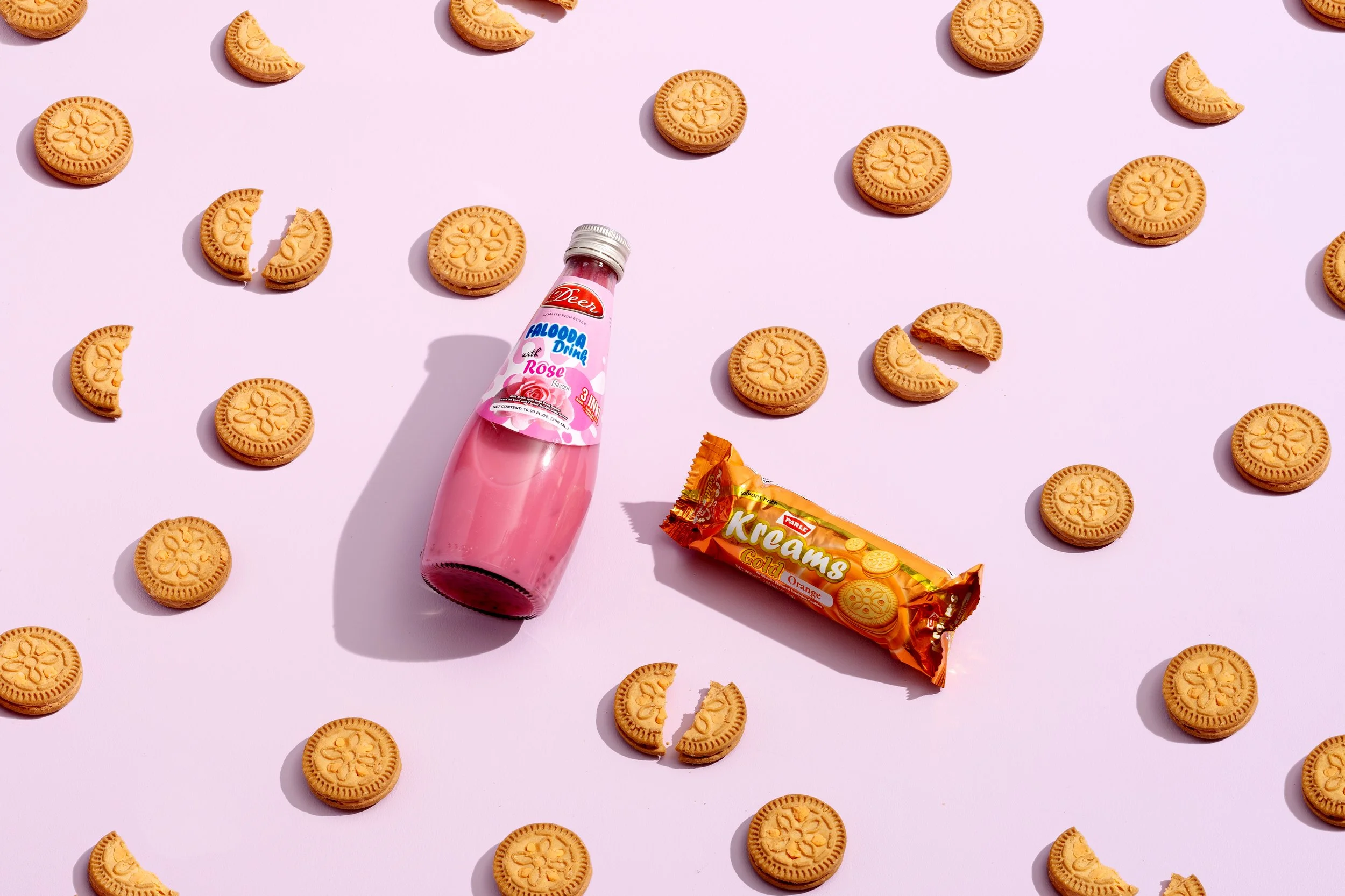 Cookies scattered on a pink background with a bottle of rose-flavored drink and a package of orange-flavored cream biscuits.