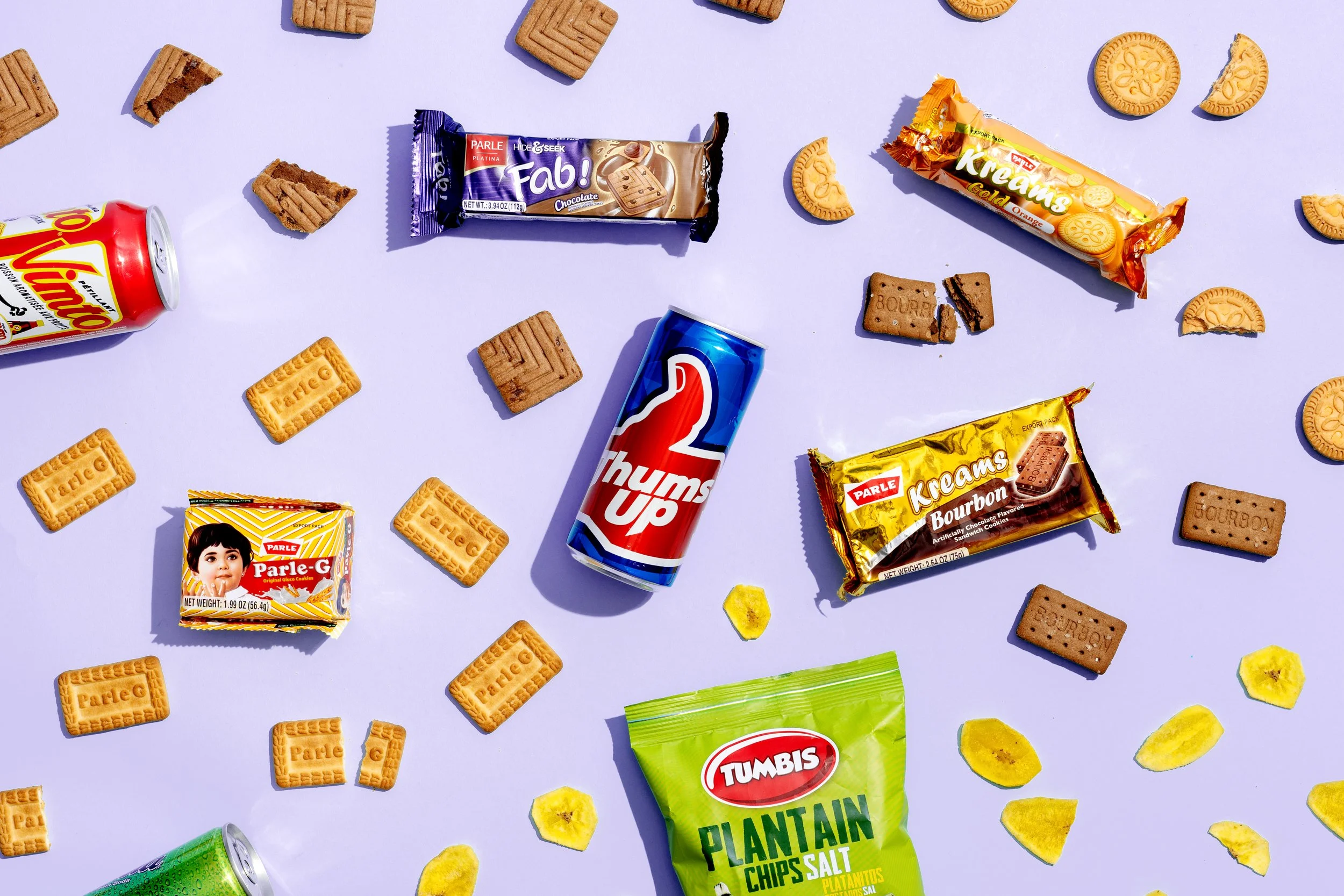 An assortment of snack items scattered on a purple background, including cookies, chips, and soda cans.