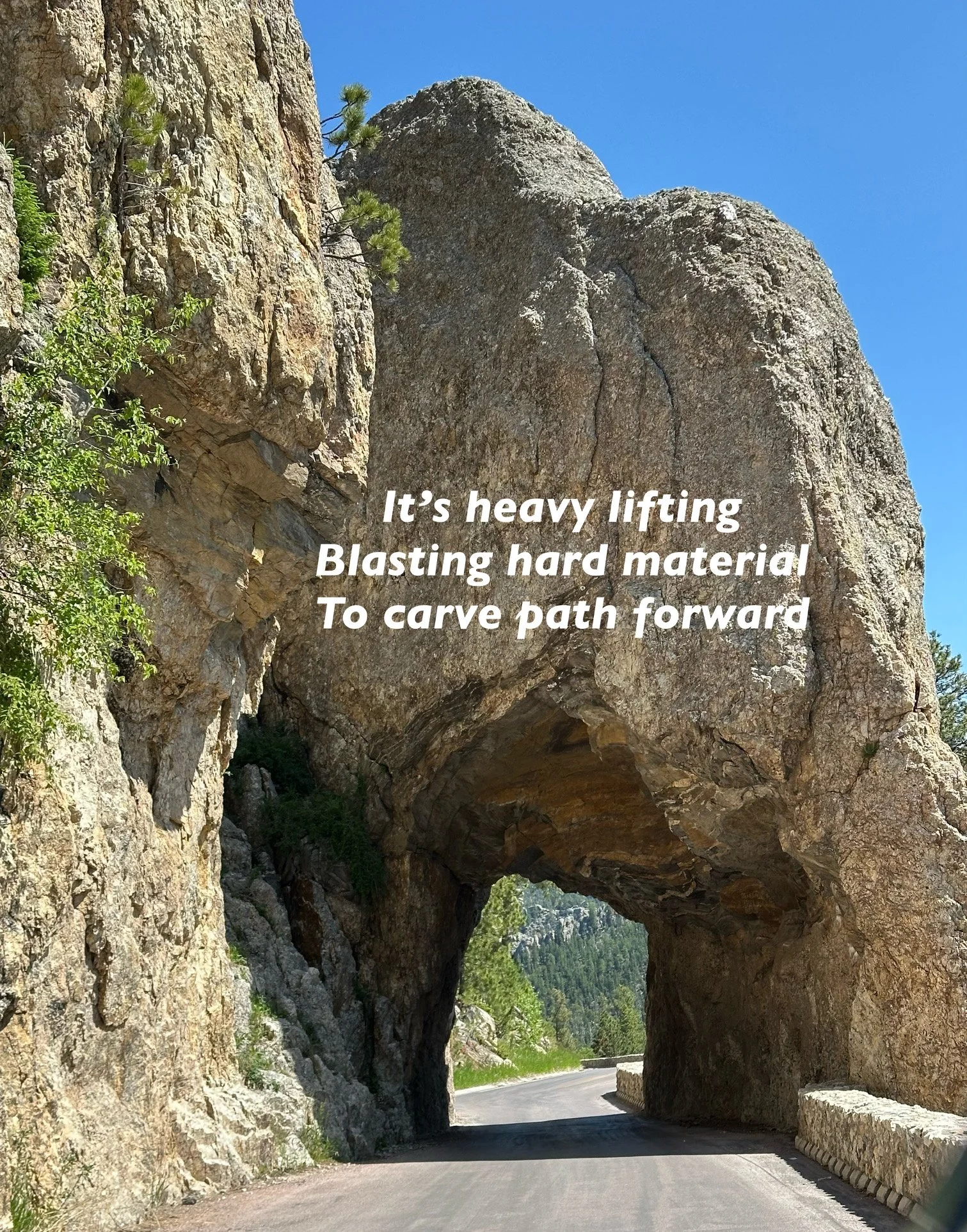 A narrow mountain road passing through a large rock formation that creates a natural tunnel. The surroundings are rocky with some greenery and a clear blue sky.