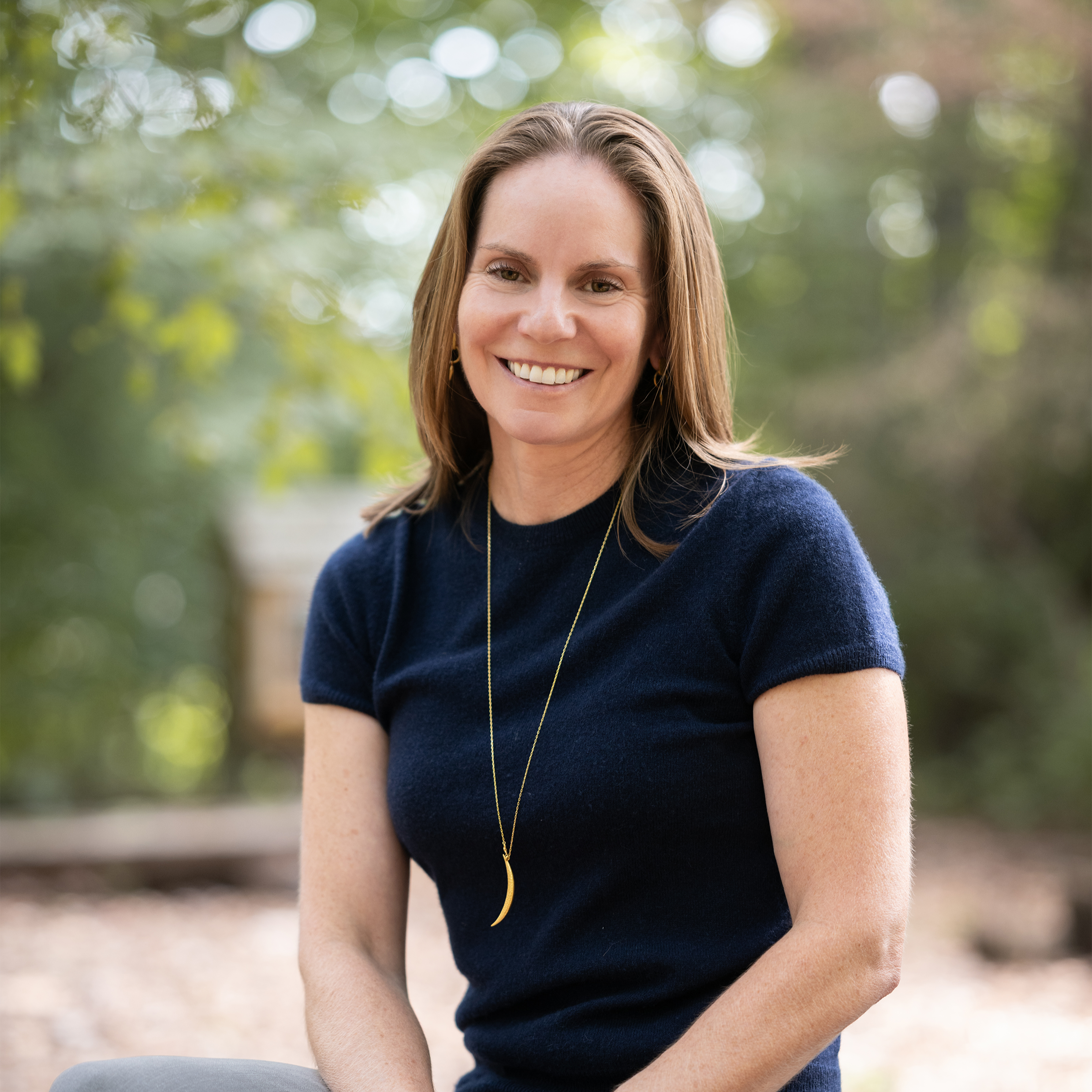 Image of Julie smiling outdoors with trees in the background, wearing a navy blue top and a gold necklace.