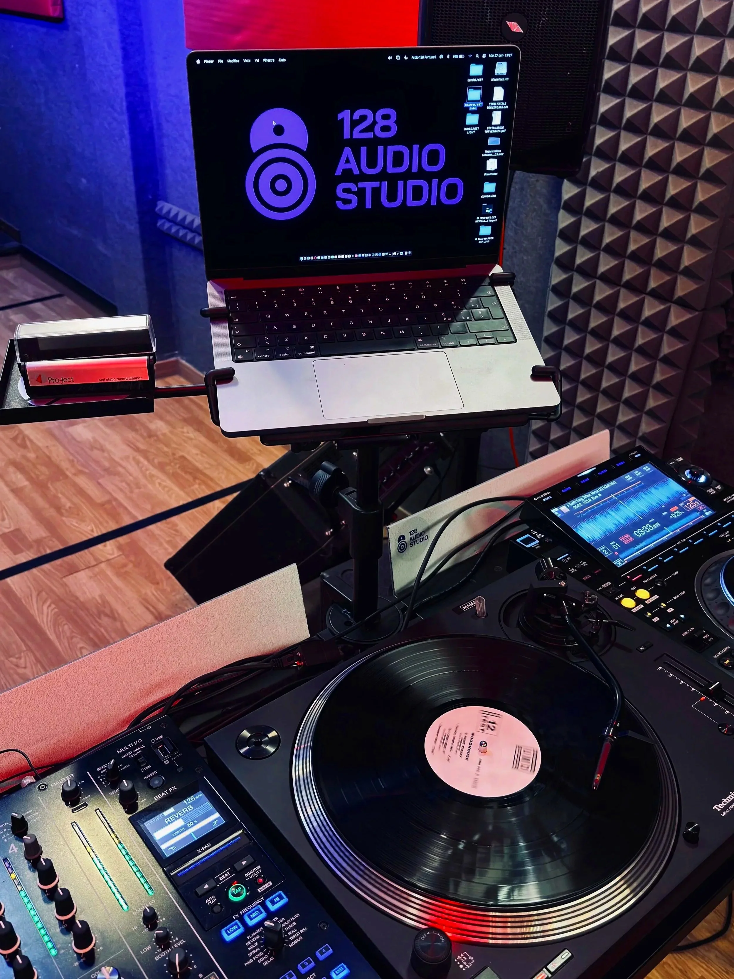 Setup di uno studio audio con un laptop su un support, un giradischi con un disco in vinile, un mixer e un controller DJ, con pannelli acustici sullo sfondo.